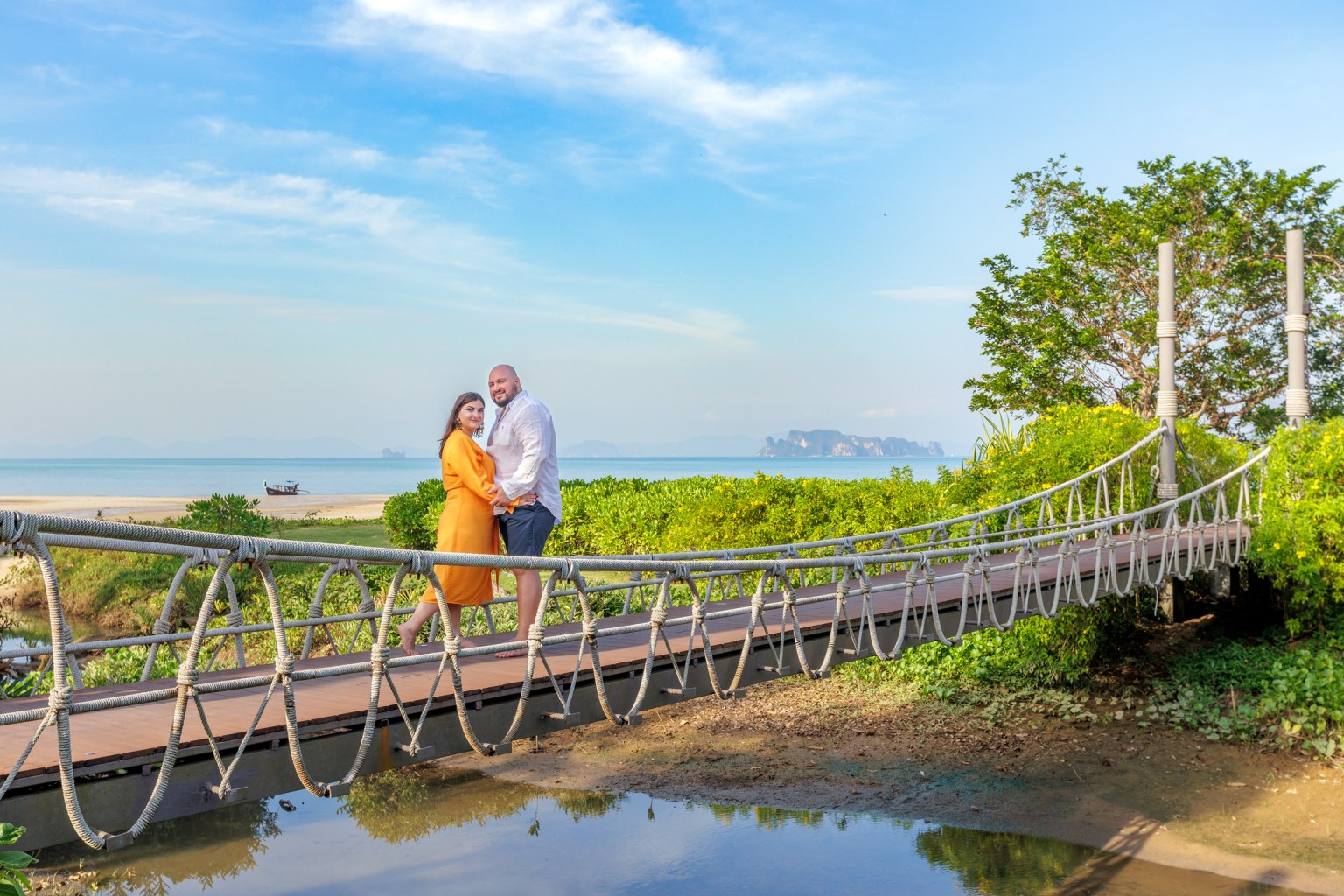 A couple stands on a rope bridge surrounded by lush greenery, with a scenic beach and distant islands in the background. The woman wears an orange dress, while the man is in a lighter shirt and shorts. The clear blue sky completes the picturesque setting.