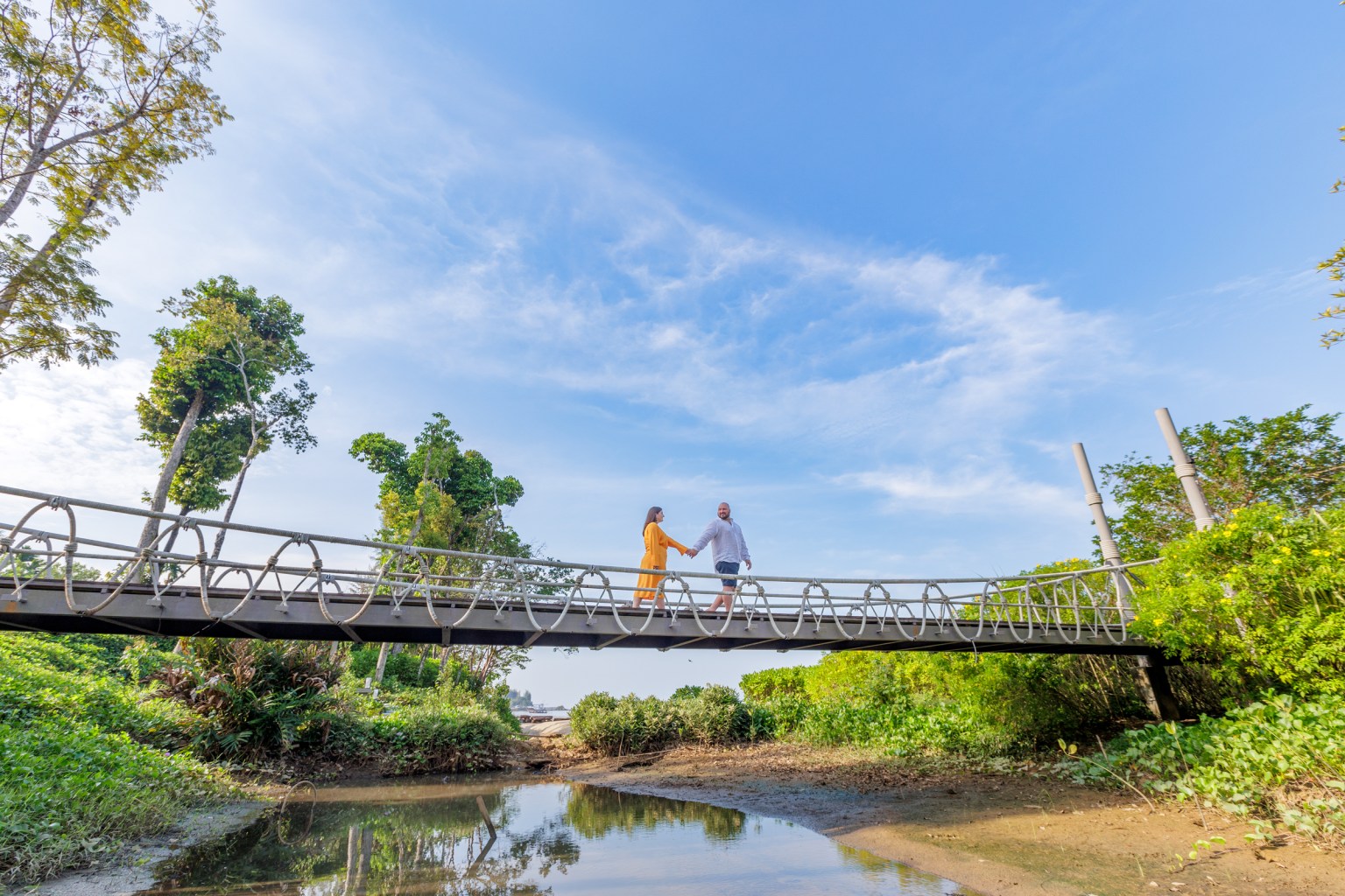 A couple holding hands on a metal bridge over a tranquil waterway surrounded by lush greenery and trees under a bright blue sky.