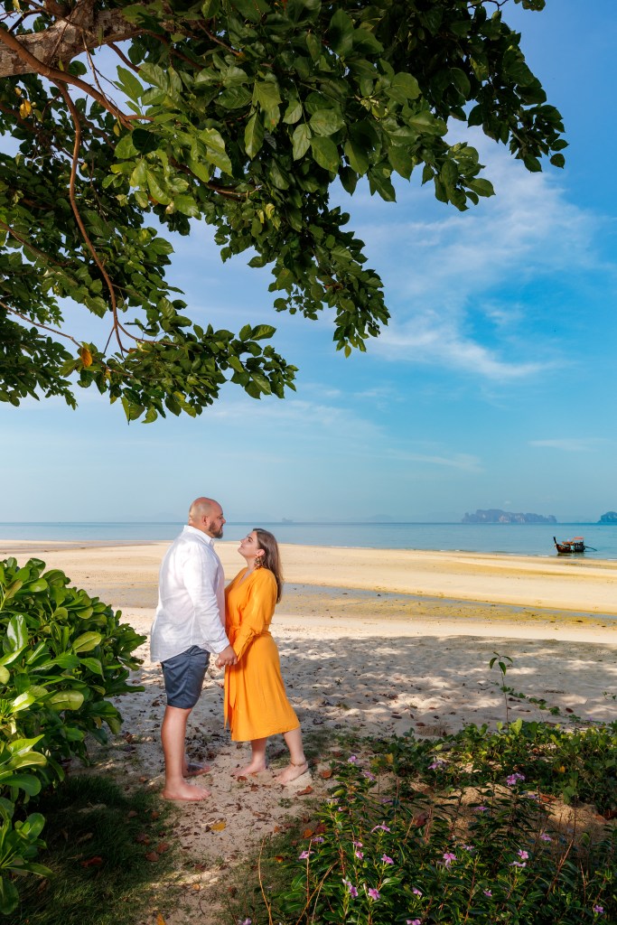 A couple gazing into each other's eyes while holding hands on a sandy beach surrounded by lush greenery and blue skies.