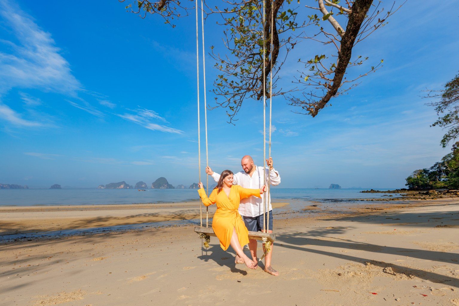 A couple enjoying a swing on a beach in Krabi, Thailand, with clear blue skies and limestone cliffs in the background.