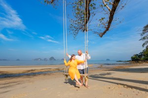 A couple enjoying a romantic moment on a swing at a beach with clear blue skies and distant islands in the background.