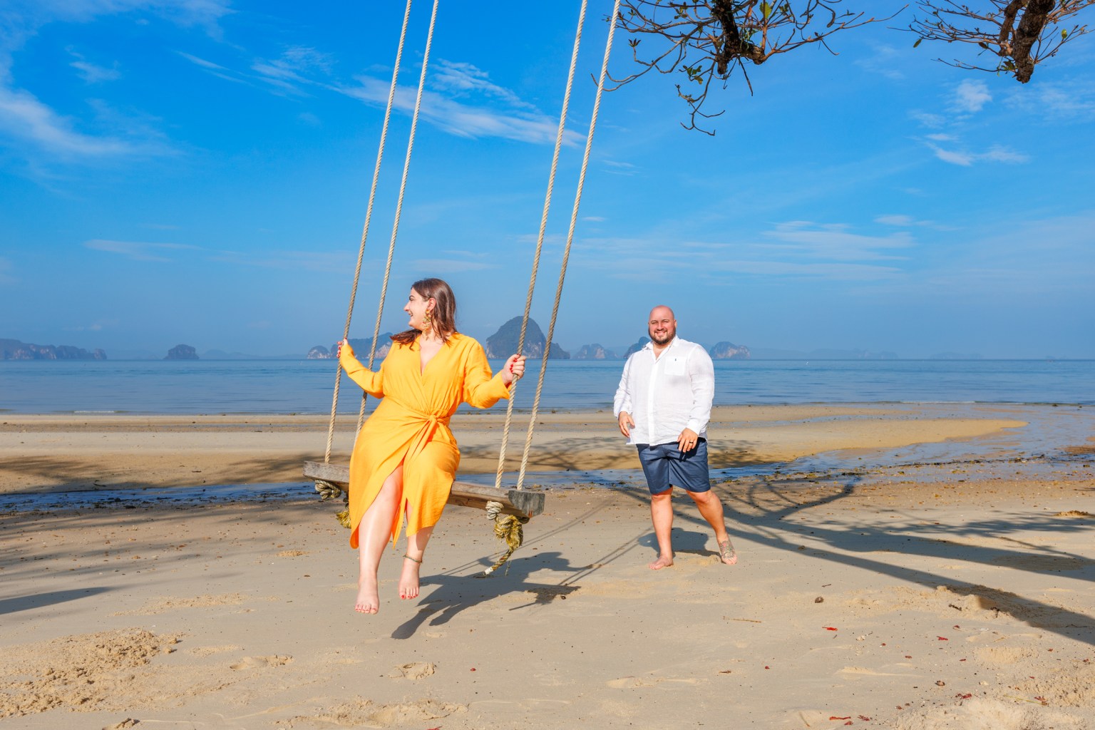 A woman in an orange dress swings on a beach swing, smiling and enjoying the view, while a man in a white shirt and shorts walks beside her on a sandy beach with the ocean in the background.
