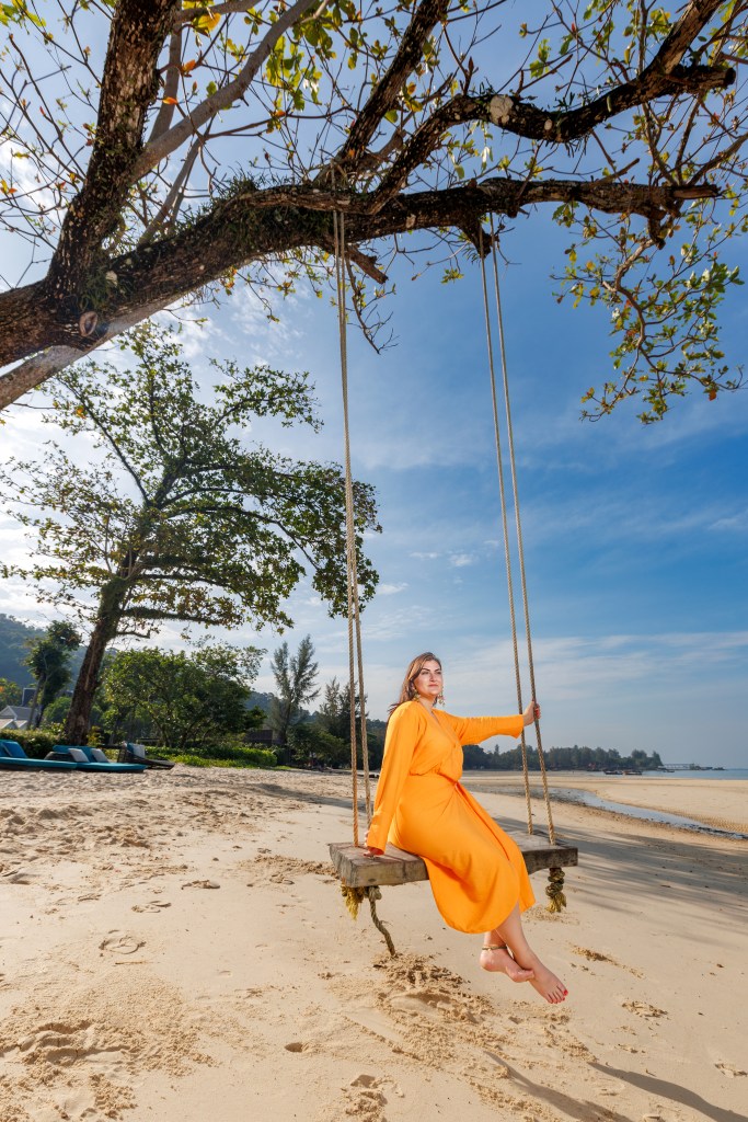 A woman in an orange dress sitting on a swing under a tree on a beach. There are beach chairs in the background and a clear blue sky.