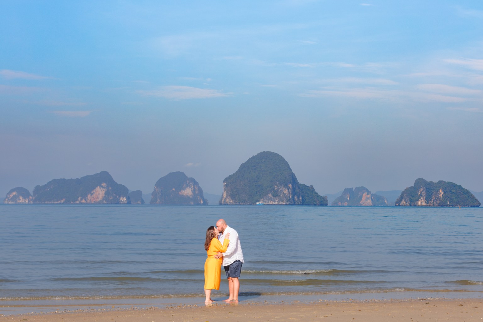 A couple sharing a kiss on the beach in Krabi, Thailand, with limestone cliffs and islands visible in the background under a clear blue sky.