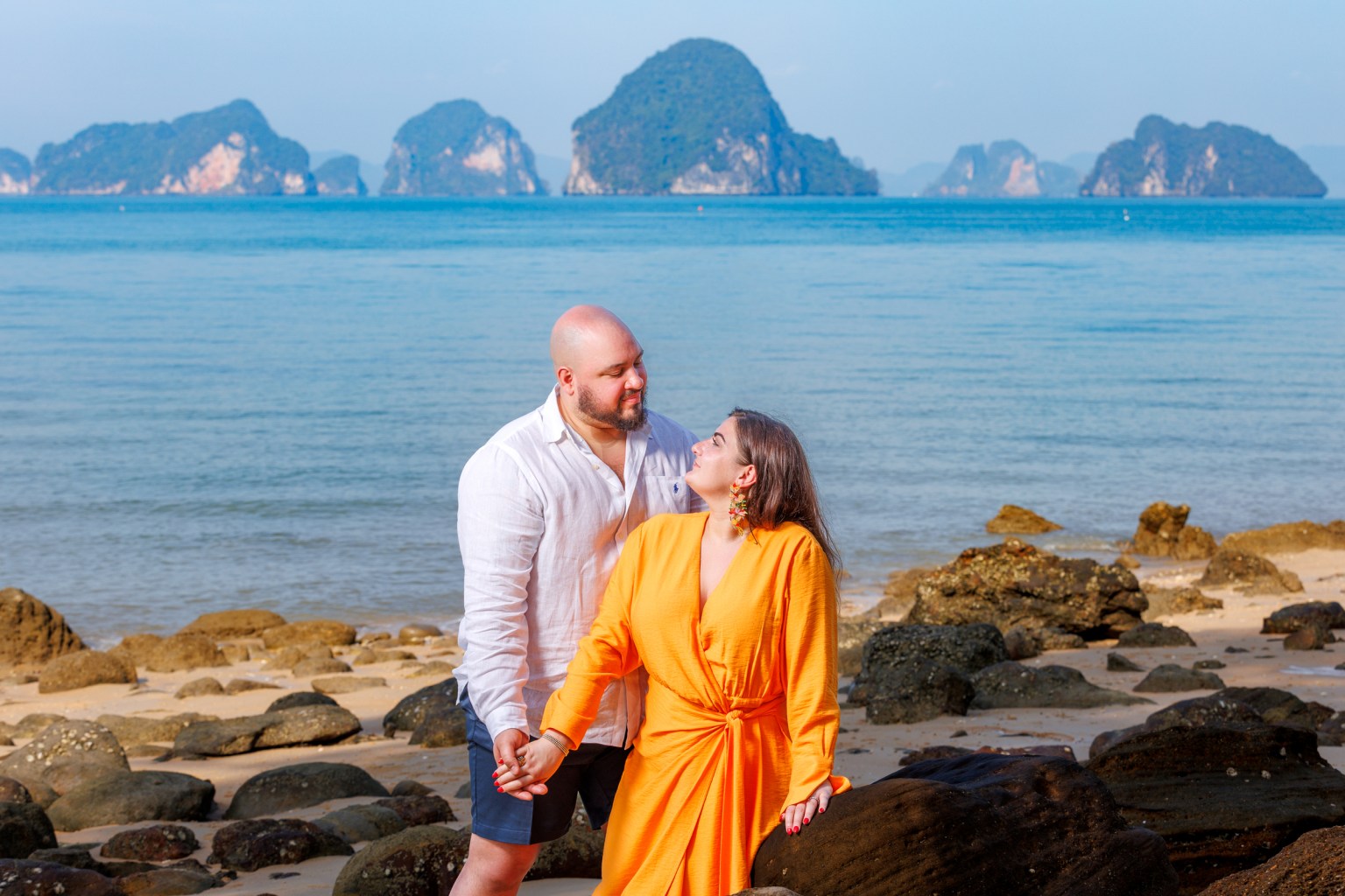 A couple stands on a rocky beach in Krabi, Thailand, gazing at each other against a backdrop of clear water and distant limestone islands.