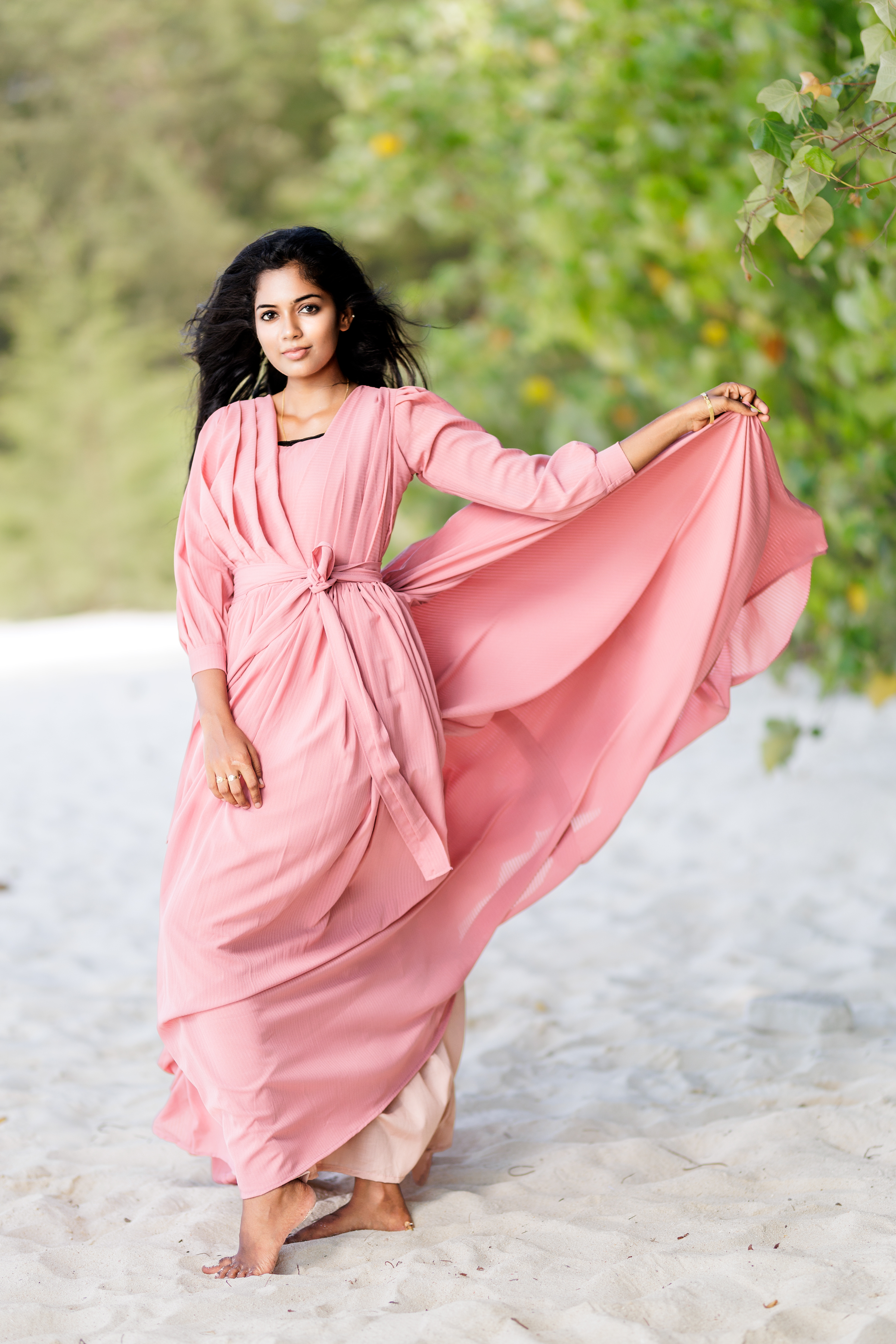 A woman in a flowing pink dress poses gracefully on a sandy beach, surrounded by lush greenery.