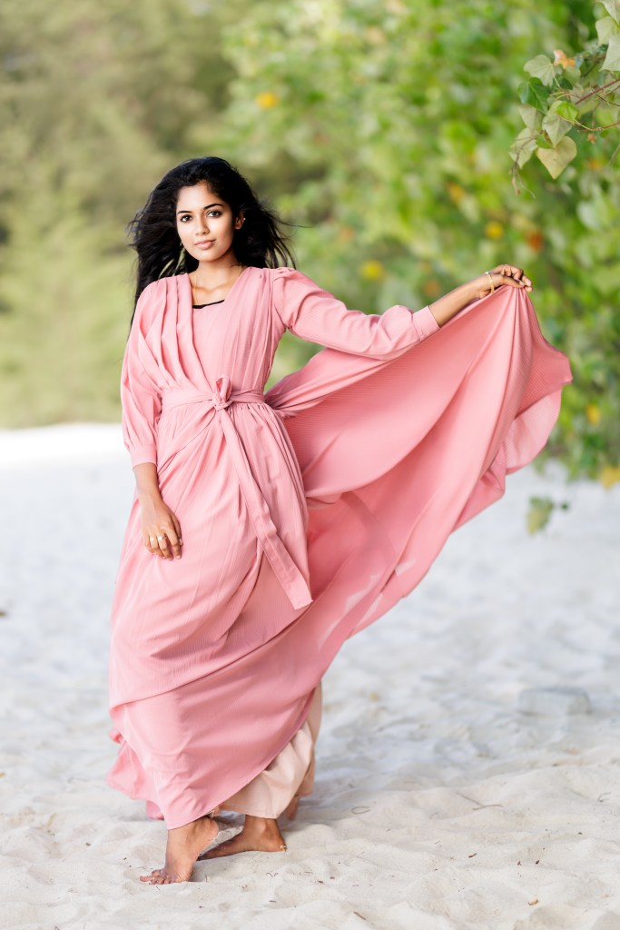 A woman wearing a pink dress with flowing fabric stands barefoot on a sandy beach, surrounded by greenery.