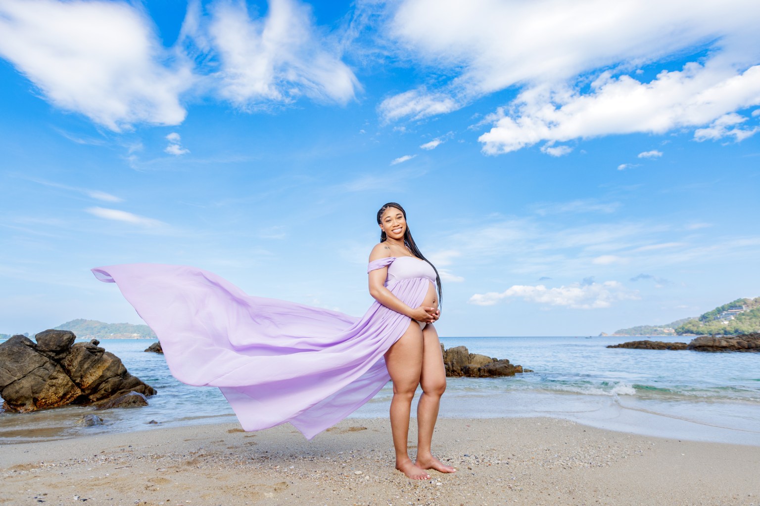 Pregnant woman in a flowing lavender dress standing on the beach, smiling with hands on her belly, against a backdrop of clear blue sky and sea.