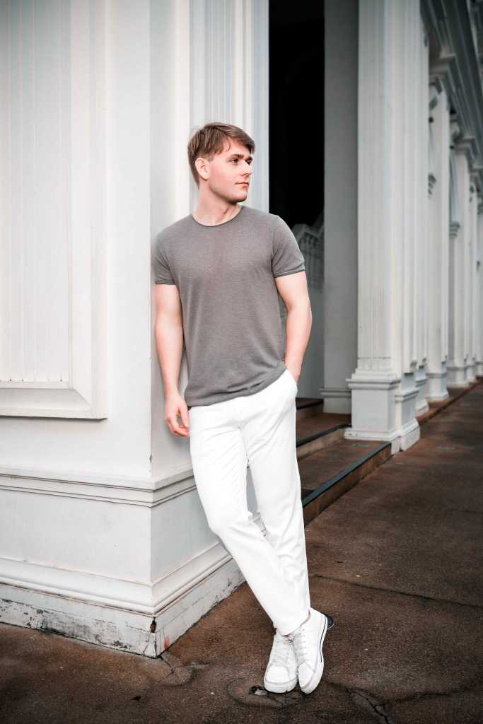 A young man in a gray shirt and white pants stands casually against a column, with a stylish urban background.