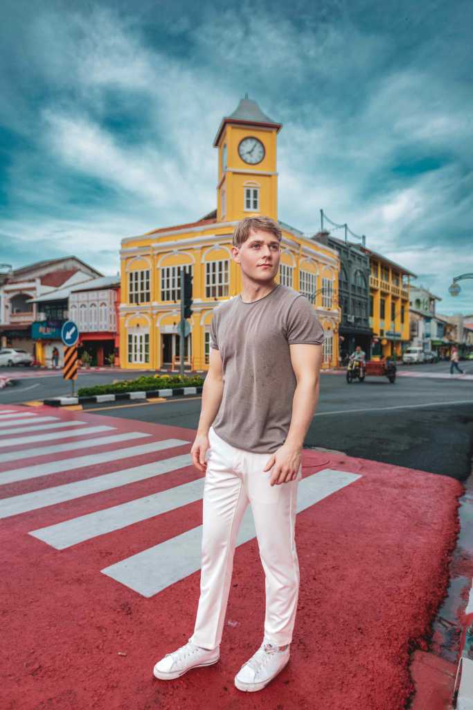 A young man stands confidently on a red crosswalk in front of a vibrant yellow building with a clock tower, against a cloudy blue sky.