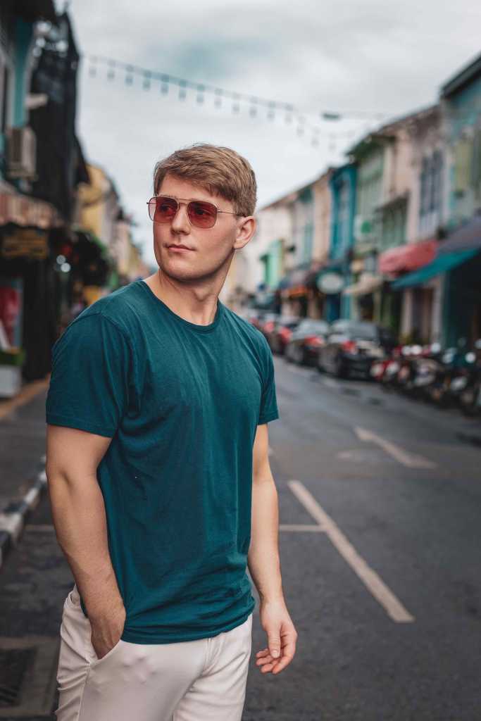 A young man wearing sunglasses and a teal t-shirt poses confidently in a vibrant street scene, with colorful buildings and parked vehicles in the background.