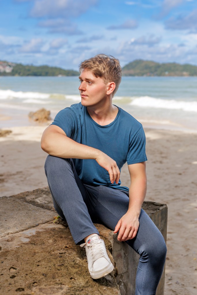 A young man seated on a rock at a beach, looking out towards the ocean, with gentle waves and a clear blue sky in the background.