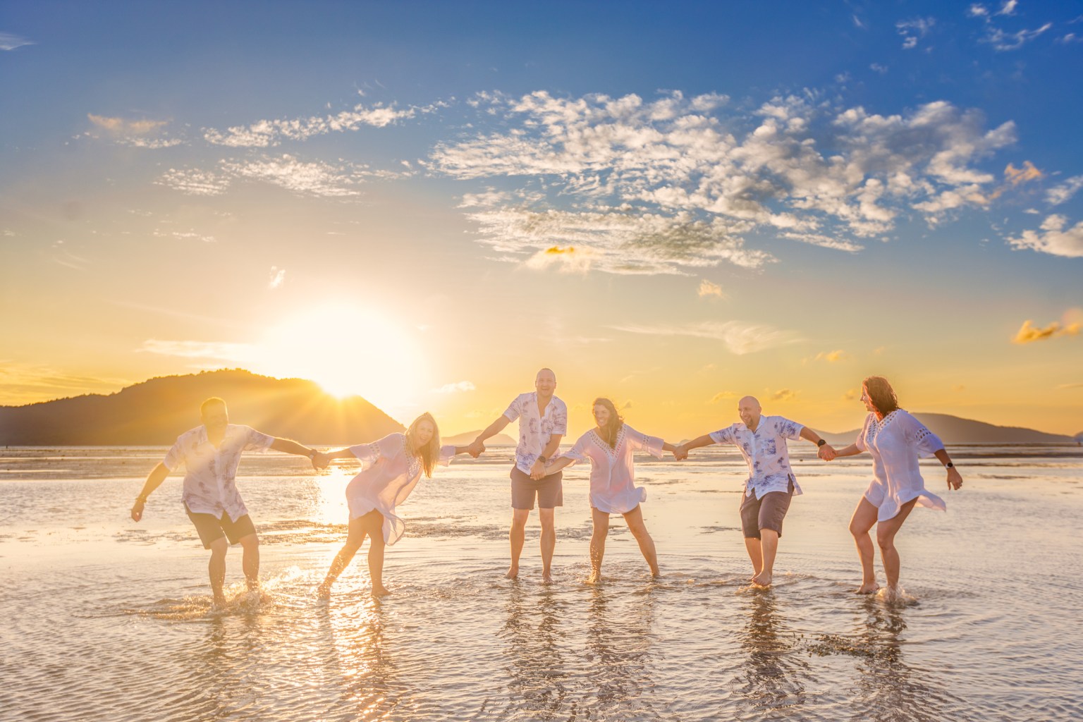 Group of six people holding hands and standing in shallow water during sunrise at the beach in Phuket, with a vibrant sky and mountains in the background.