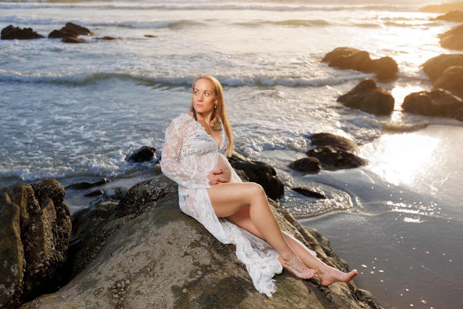 A pregnant woman in a lace dress sitting on a rock by the beach, gazing thoughtfully at the ocean during sunset.