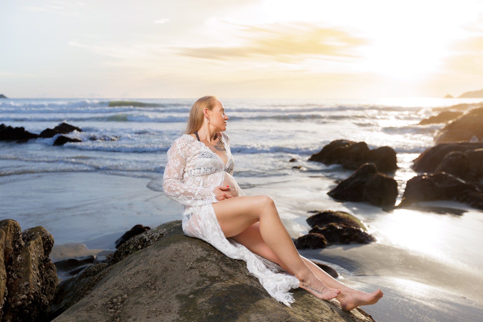 A pregnant woman wearing a lace dress sits gracefully on a rock by the beach, gazing towards the ocean as the sun sets in the background.