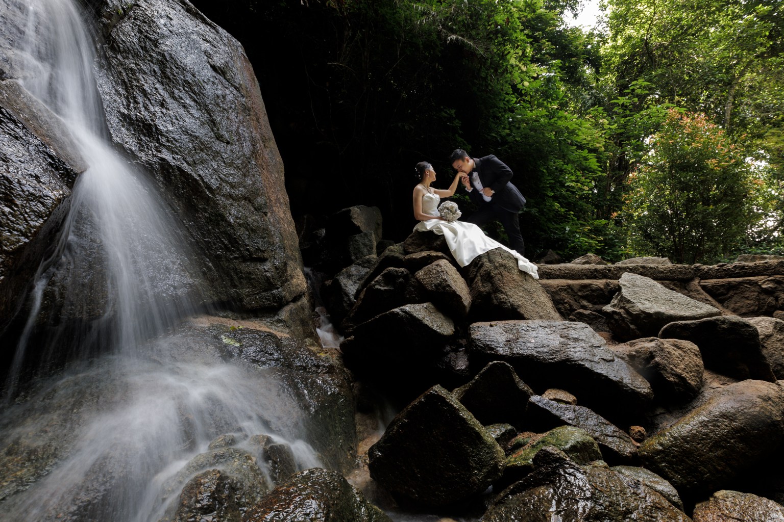 A bride and groom pose romantically on rocks near a waterfall, surrounded by lush greenery.