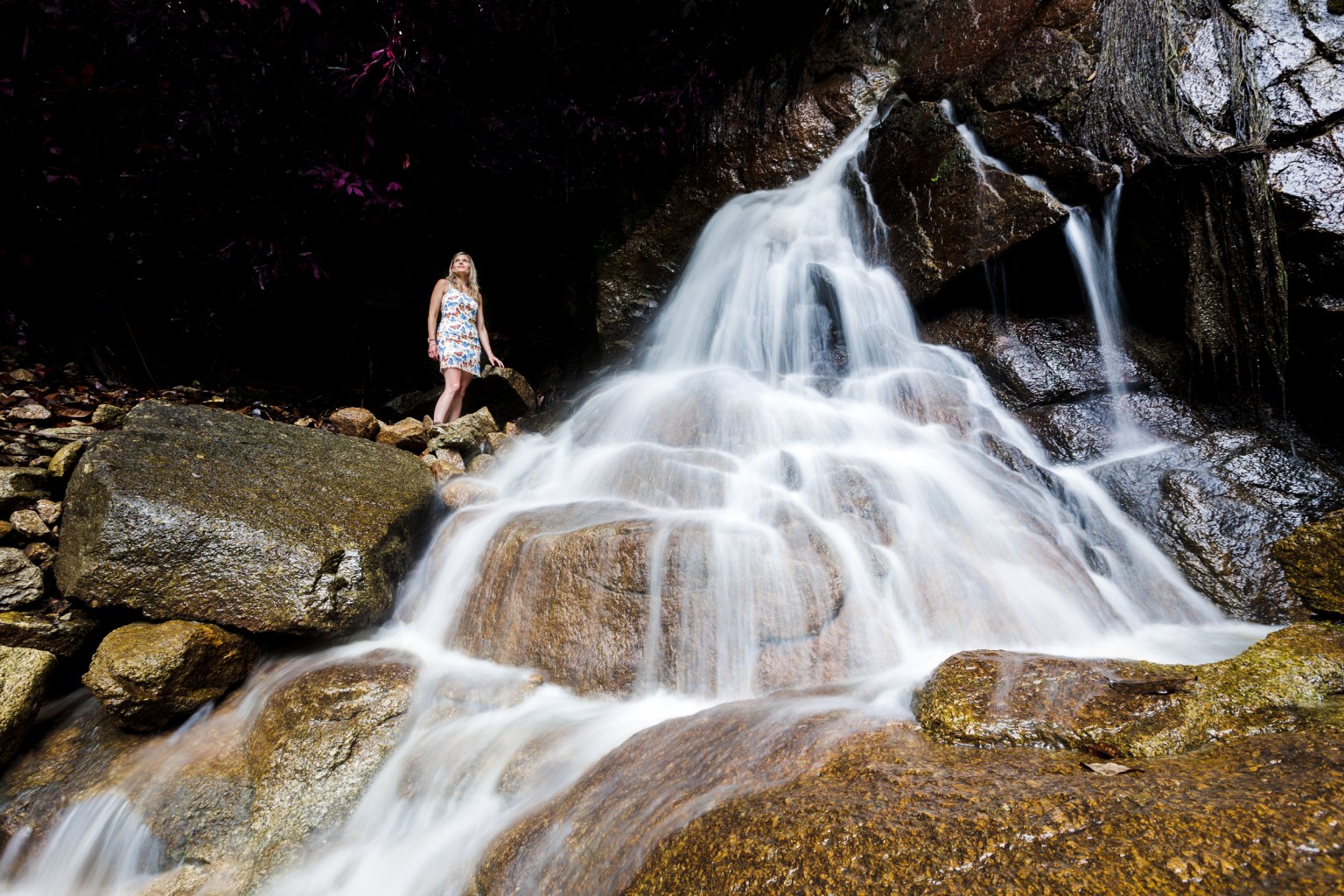 A woman standing near a waterfall, surrounded by rocks and lush greenery.