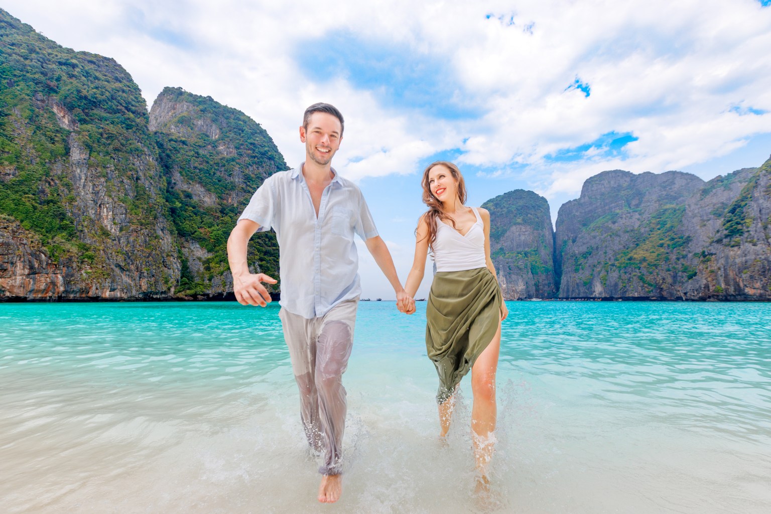 A couple joyfully running hand in hand through the shallow waters of Maya Bay, with lush limestone cliffs and a clear blue sky in the background.