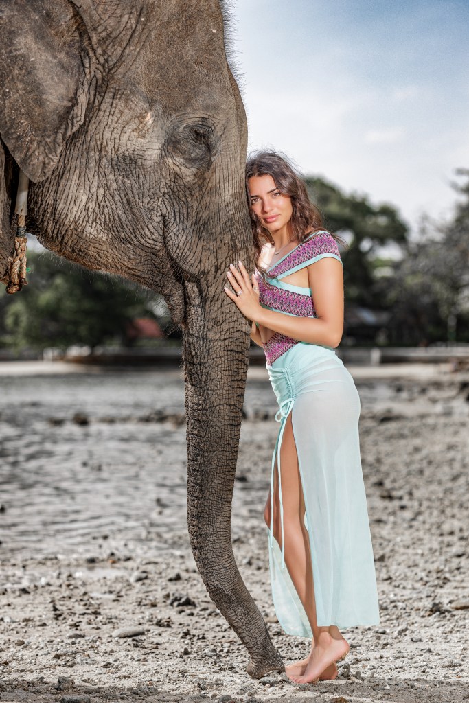 A woman poses affectionately with an elephant by her side on a sandy beach, wearing a colorful outfit. The background features lush greenery and a clear sky.