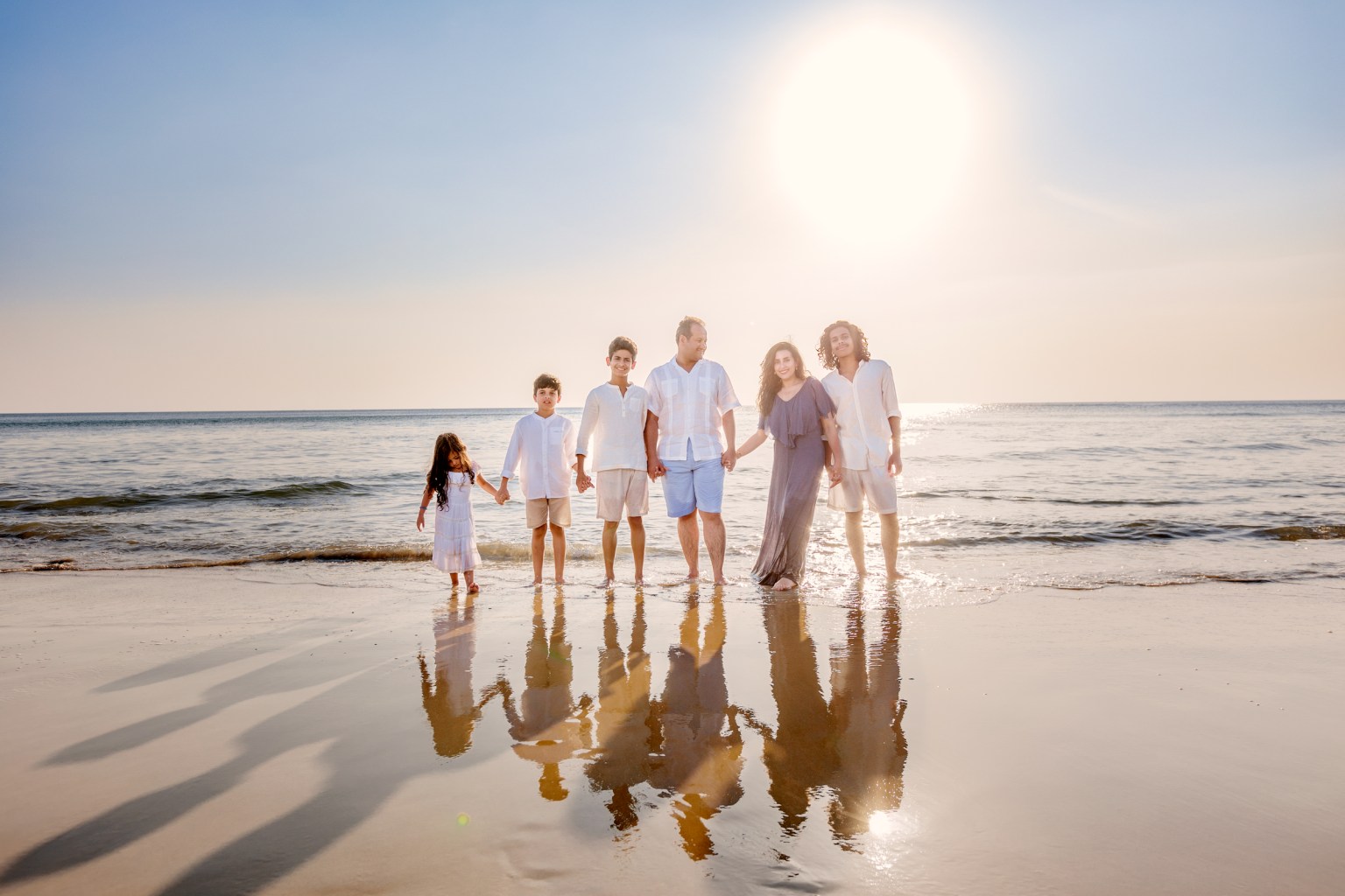 A family of six stands on a beach, holding hands with the ocean in the background and the sun setting, casting reflections in the water.