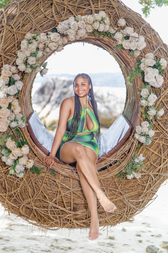 A woman sitting on a decorative circular swing adorned with flowers, wearing a green patterned swimsuit, on a beach with a scenic background.