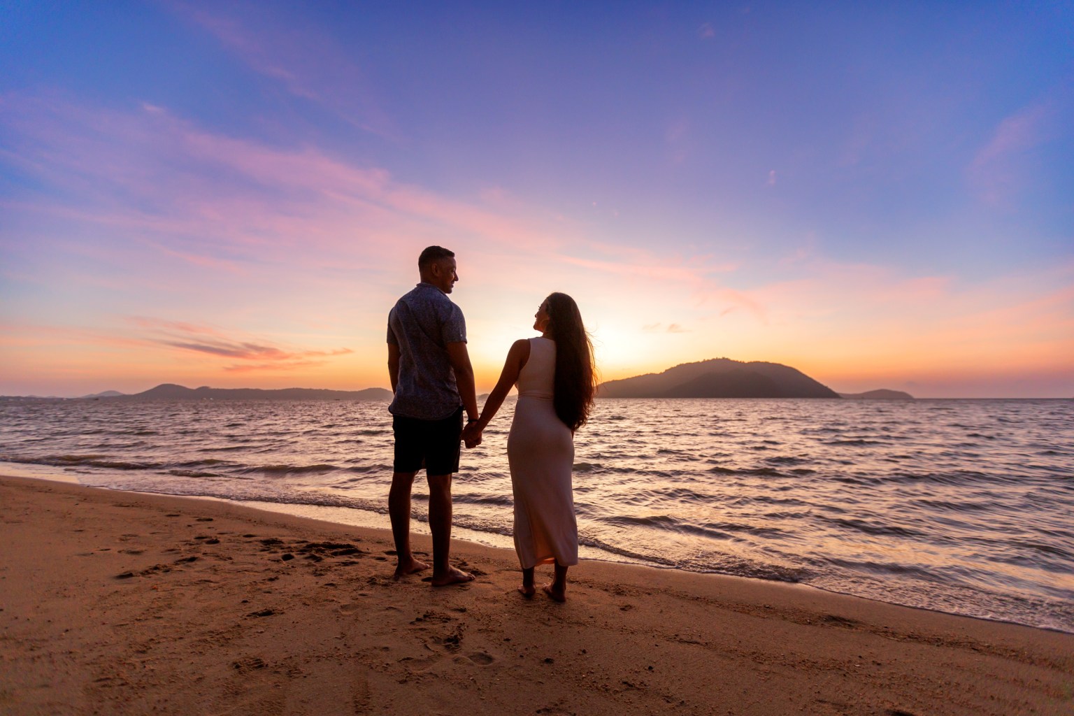 A couple holding hands on a beach at sunrise , with waves lapping at the shore and a colorful sky in the background.