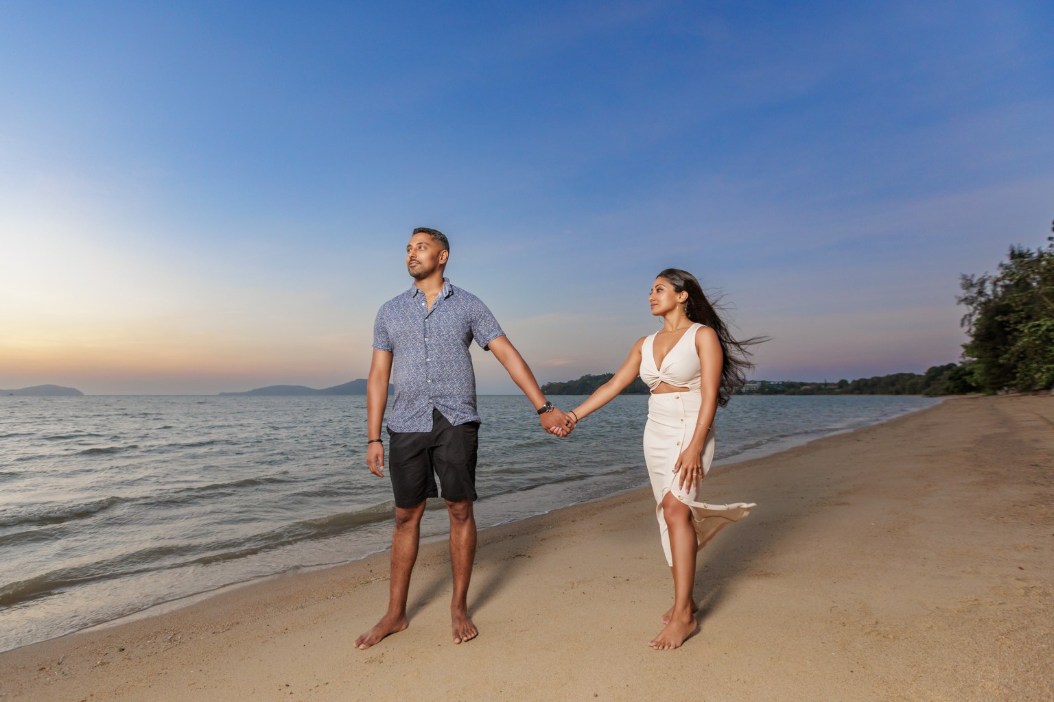 A couple holding hands on a beach during sunrise, with the ocean and distant hills in the background.