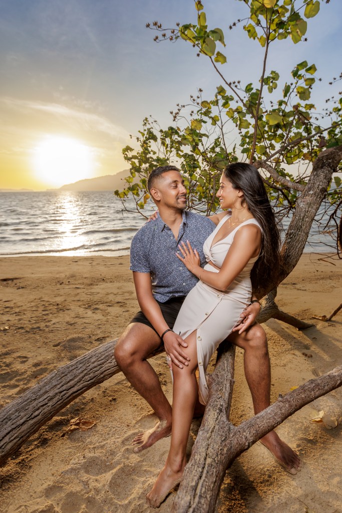 A couple sitting on a tree branch at the beach during sunrise, smiling at each other.