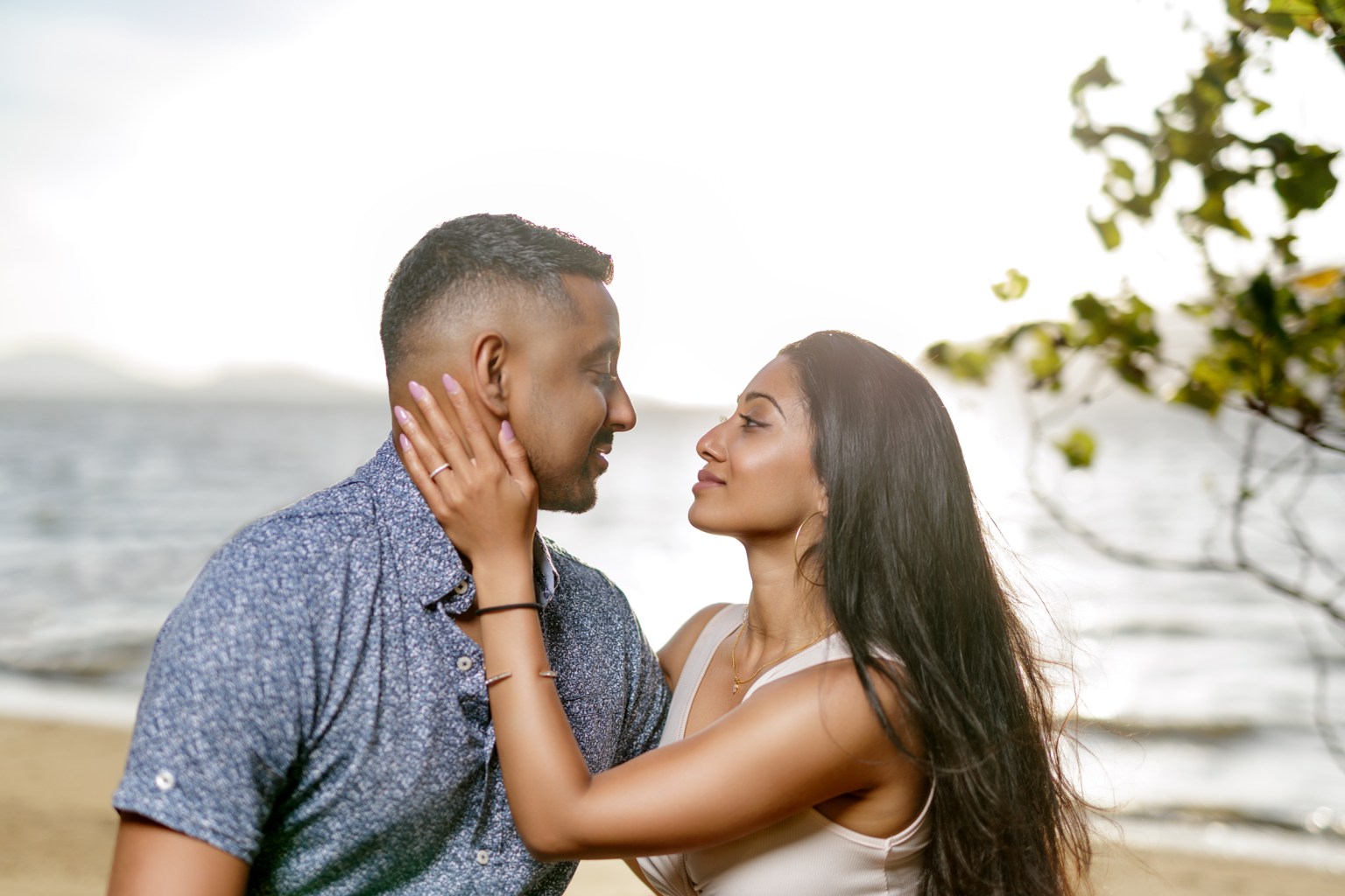 A romantic couple gazes into each other's eyes on a beach, with the ocean in the background and soft sunlight illuminating the scene.
