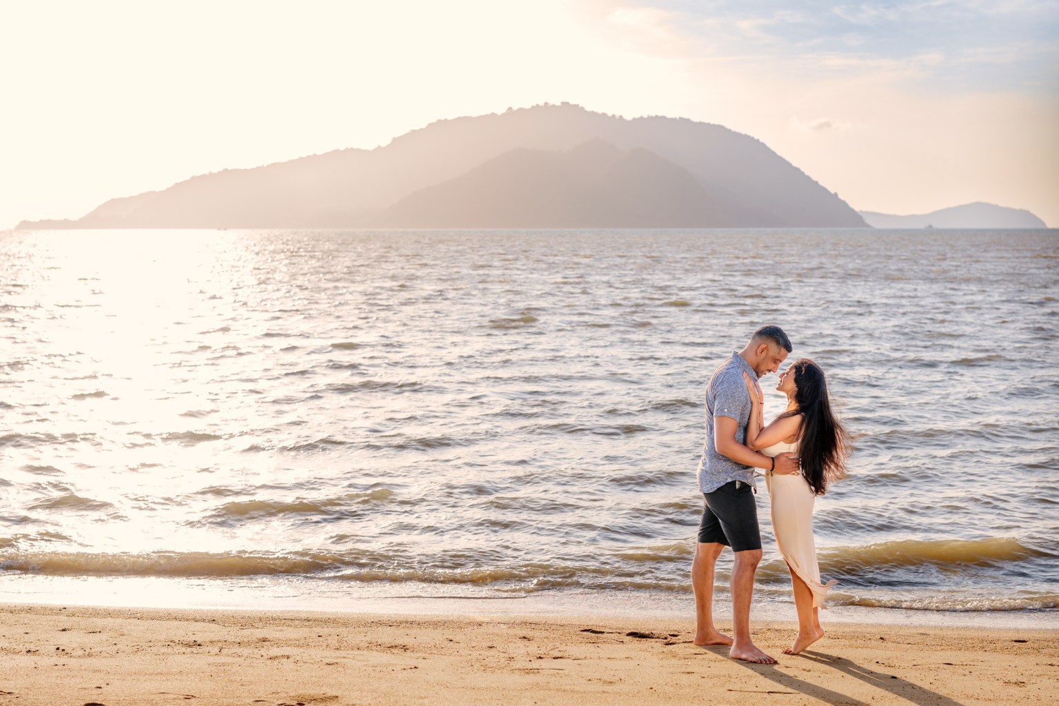 A couple embracing on a beach during sunrise, with gentle waves lapping at the shore and mountains in the background.