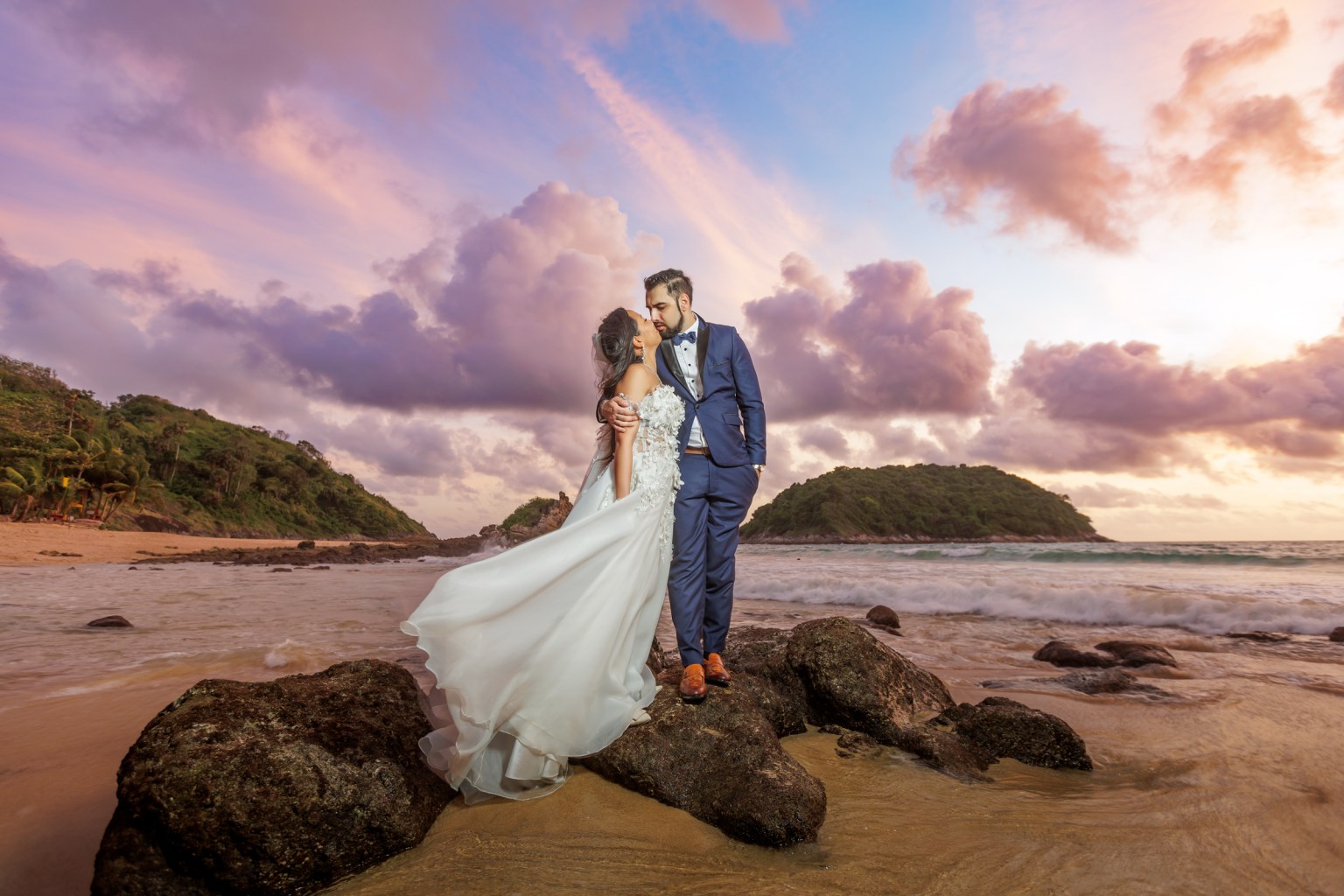 Couple in wedding attire embracing on rocks at Ya Nui Beach during sunset, with colorful clouds in the sky.