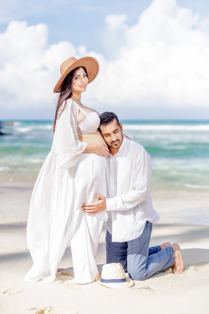 A couple on a beach during a maternity photoshoot. The woman is wearing a white flowing dress and a wide-brimmed hat, while the man kneels beside her, gently holding her belly.