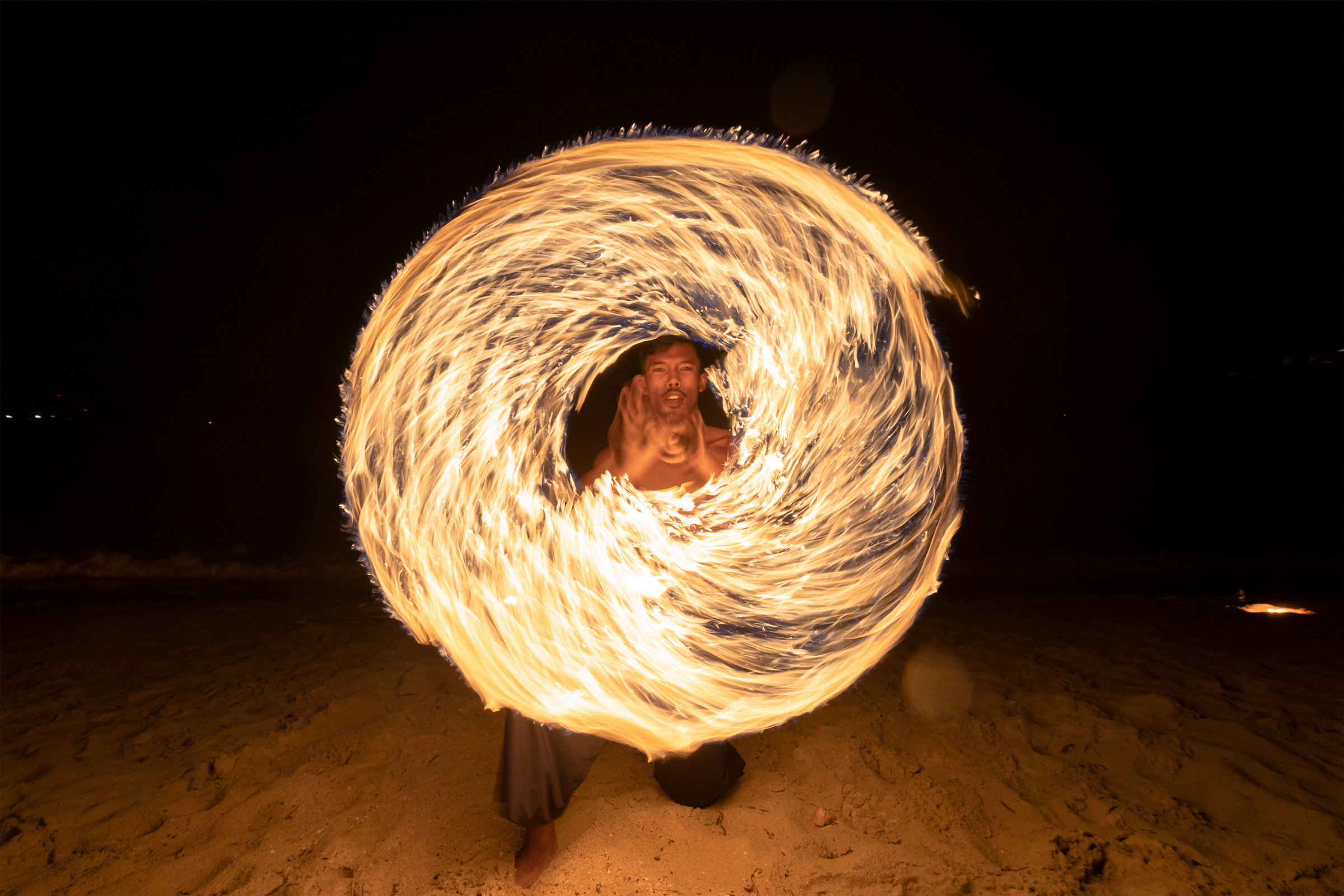 A fire performer creates a swirling ring of fire on the beach at night.