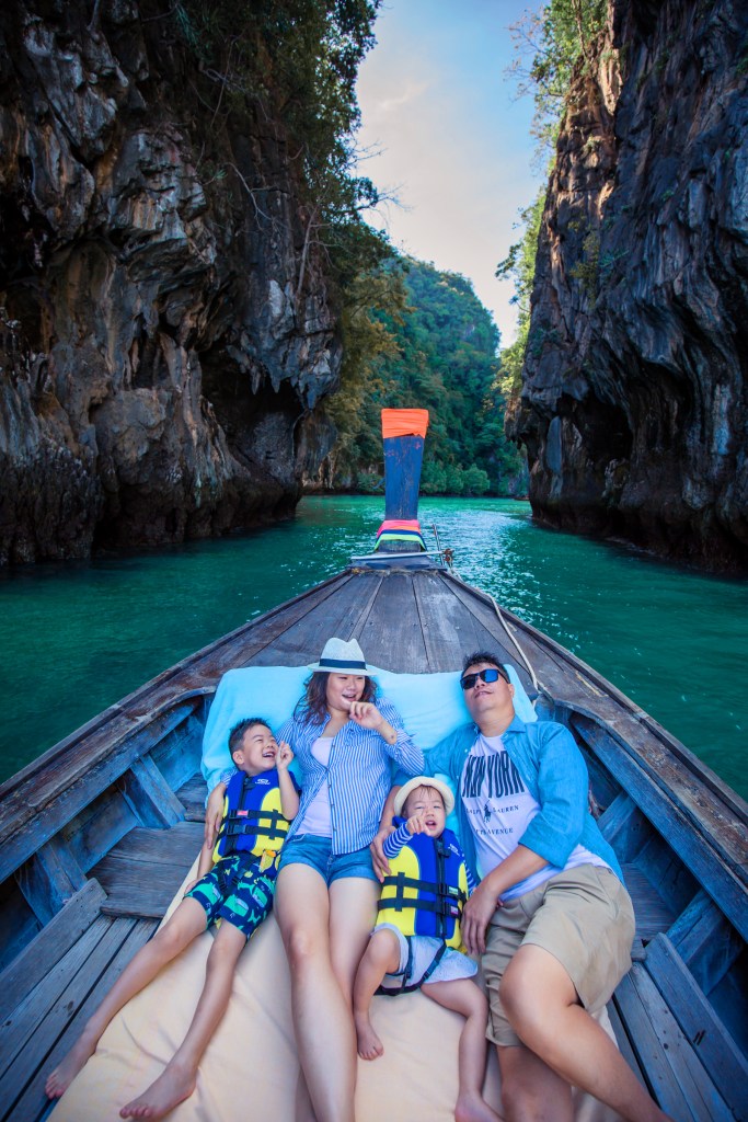 A family enjoying a boat ride in Krabi, Thailand, surrounded by towering limestone cliffs and lush greenery. Two children are playfully lying on a blanket while the adults relax beside them.
