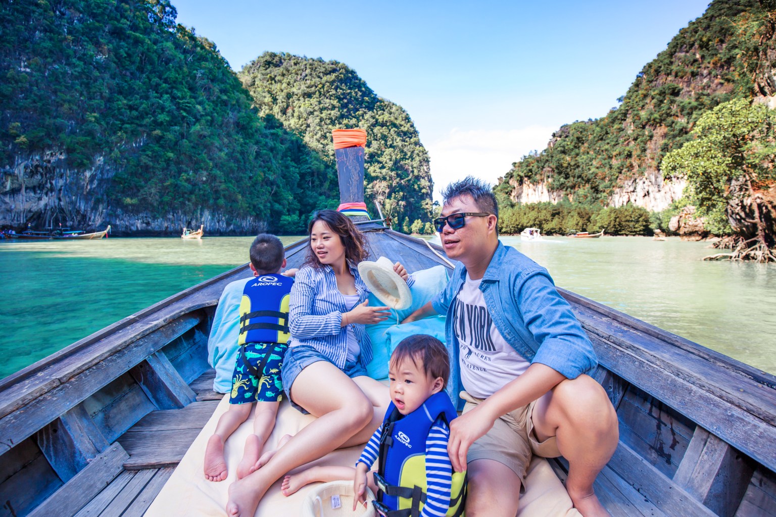 A family enjoying a boat ride in a scenic bay in Krabi, Thailand, surrounded by lush green cliffs and clear water.
