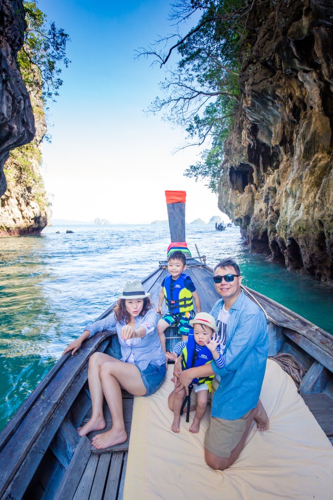 A family in a long-tail boat in Krabi, Thailand, with lush limestone cliffs in the background and a clear blue sky.