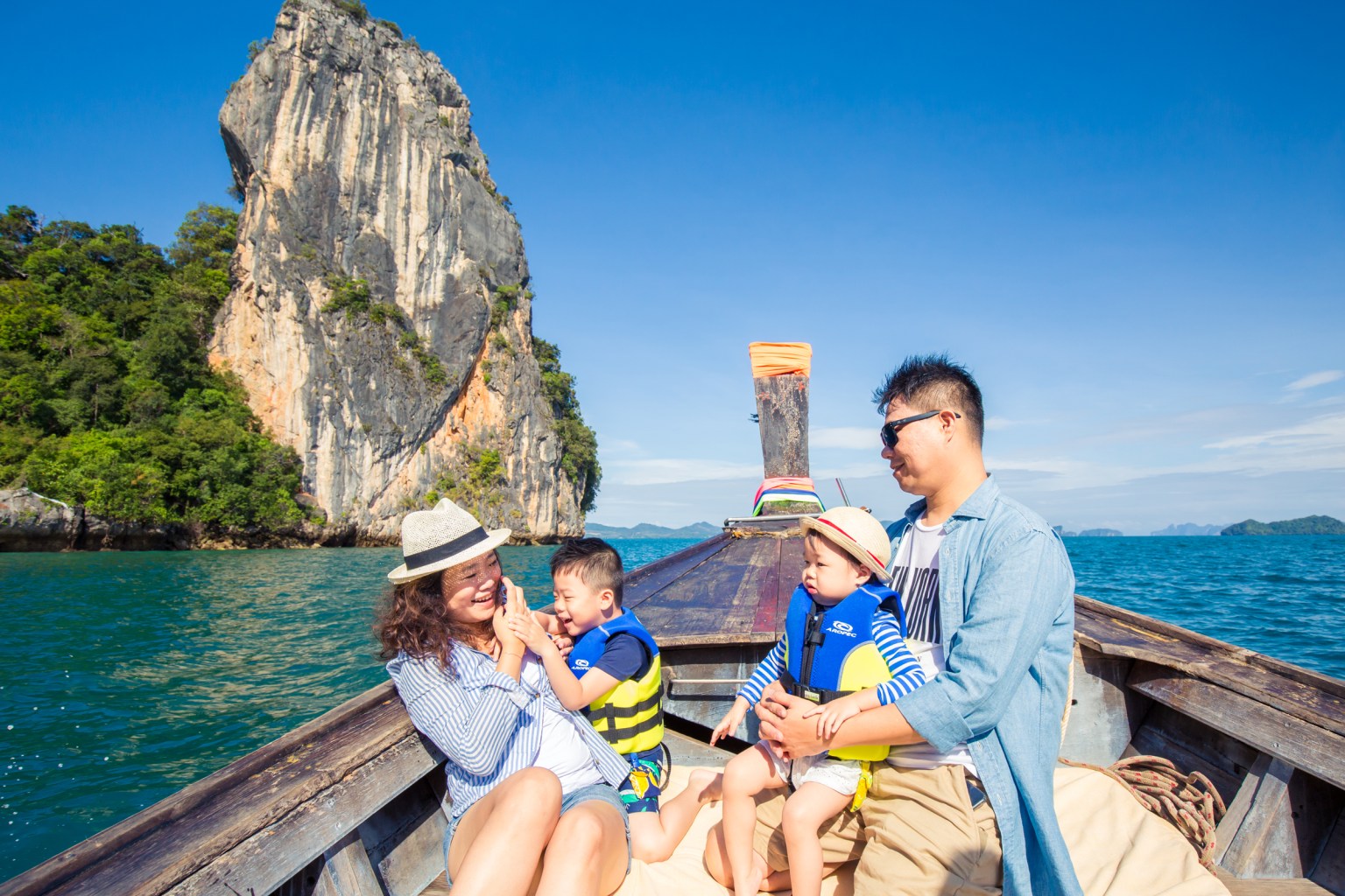 A family enjoying a boat ride in Krabi, Thailand, with a limestone cliff in the background and clear blue water.