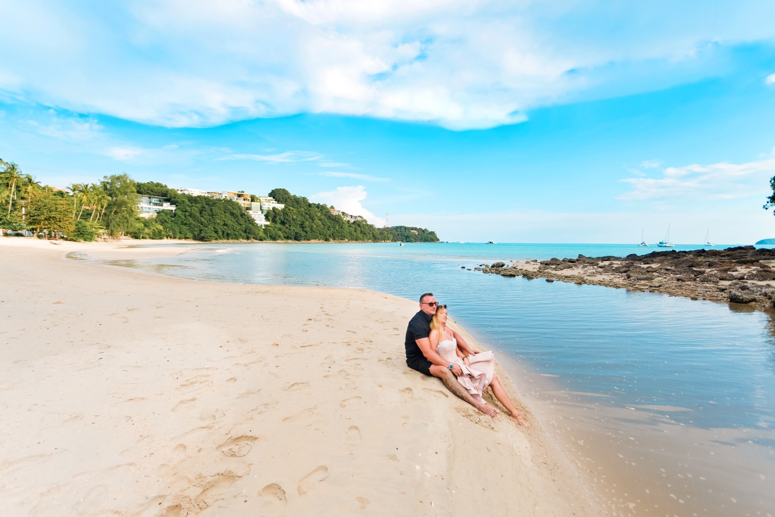 A couple sitting together on a sandy beach with calm waters and a clear blue sky.