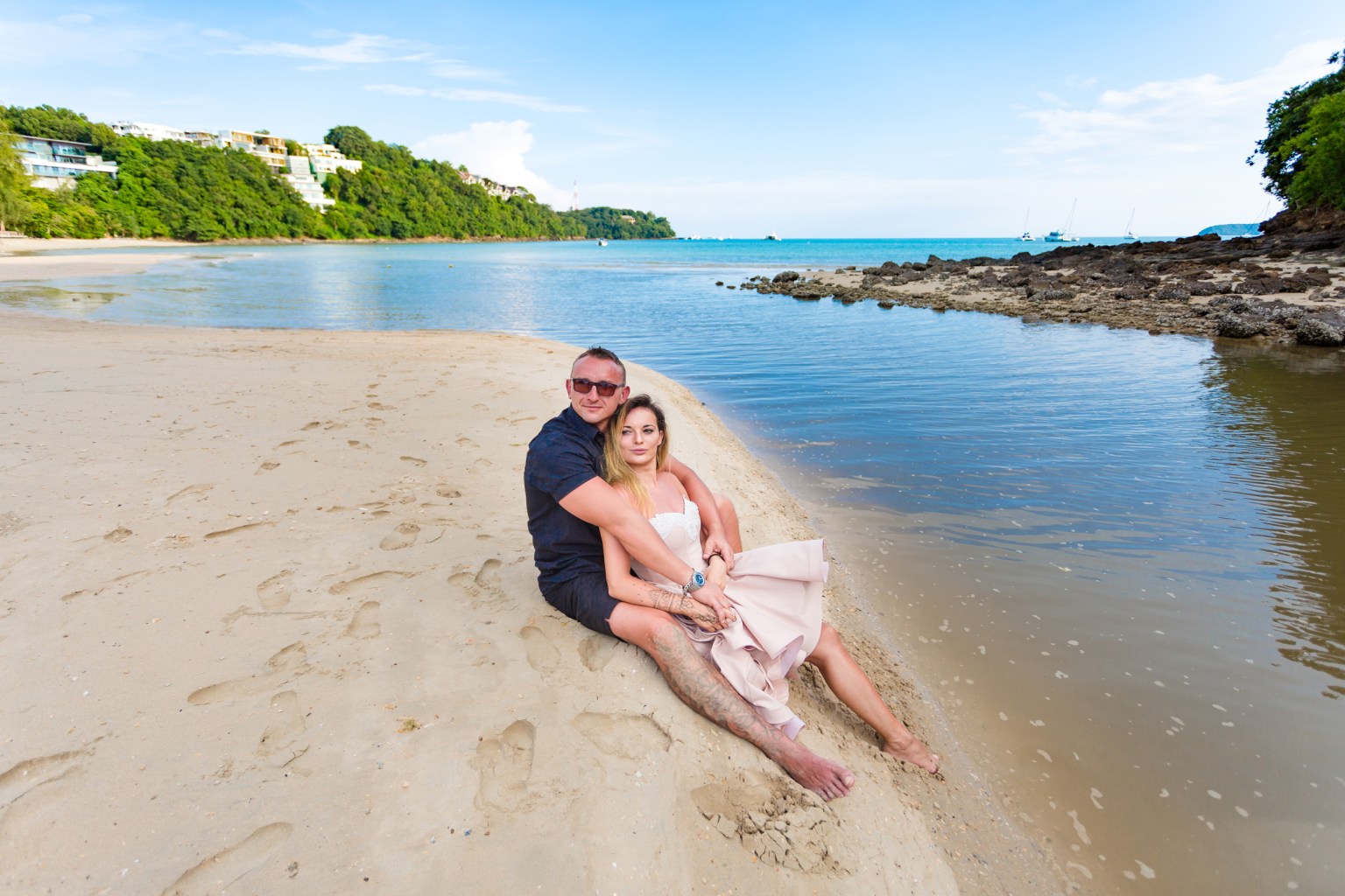 A couple sitting on the sandy beach next to a calm body of water, surrounded by greenery and rocks in the background.