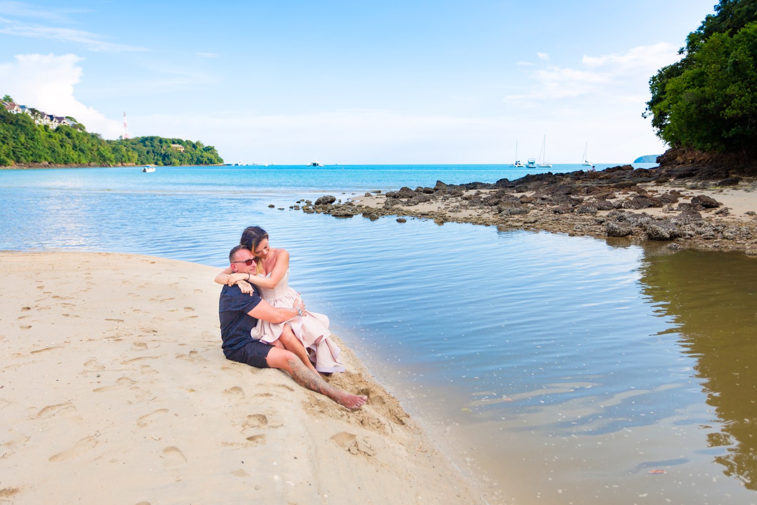 A couple embracing on a sandy beach beside calm waters and green foliage in the background.