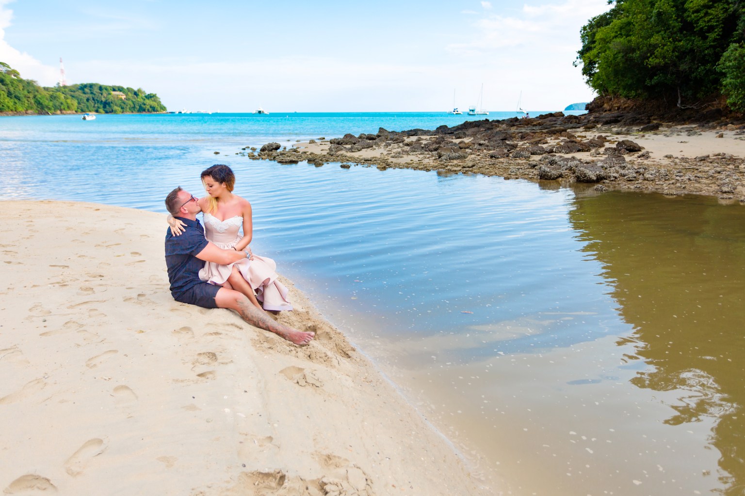 A couple sitting on the beach by a tranquil water body, with the woman in a light dress and the man in casual attire. The background features blue skies and greenery.