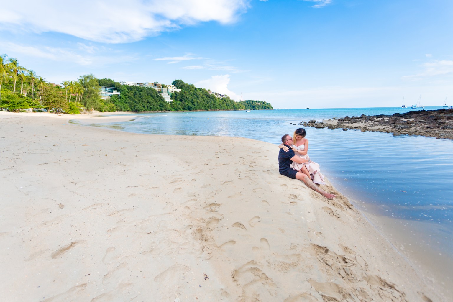 A couple sitting on the sandy beach near the water, enjoying a romantic moment with a picturesque coastal view.
