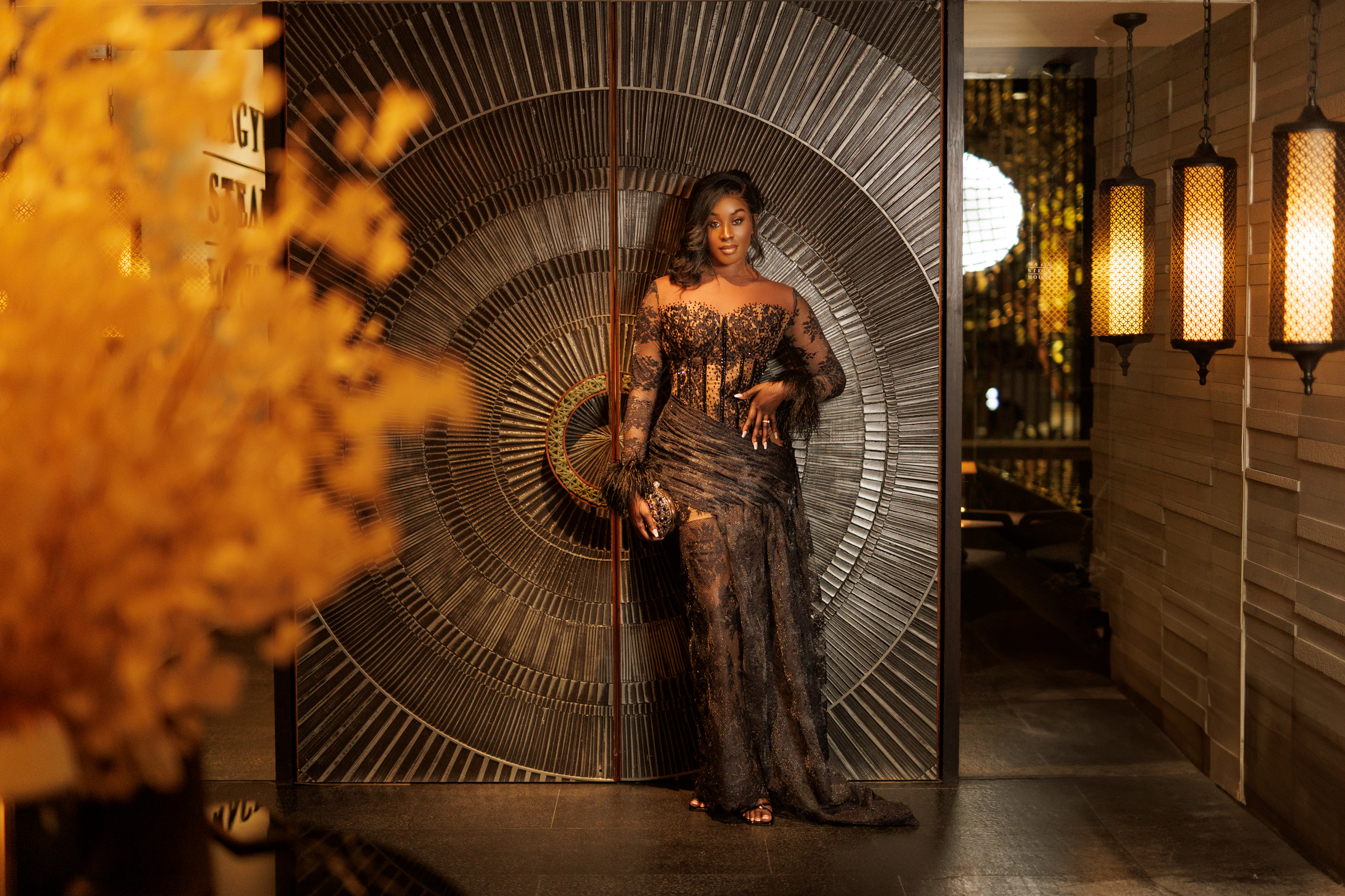 A woman in an elegant black dress stands in front of a decorative wall with a circular pattern, showcasing a sophisticated fashion portrait.