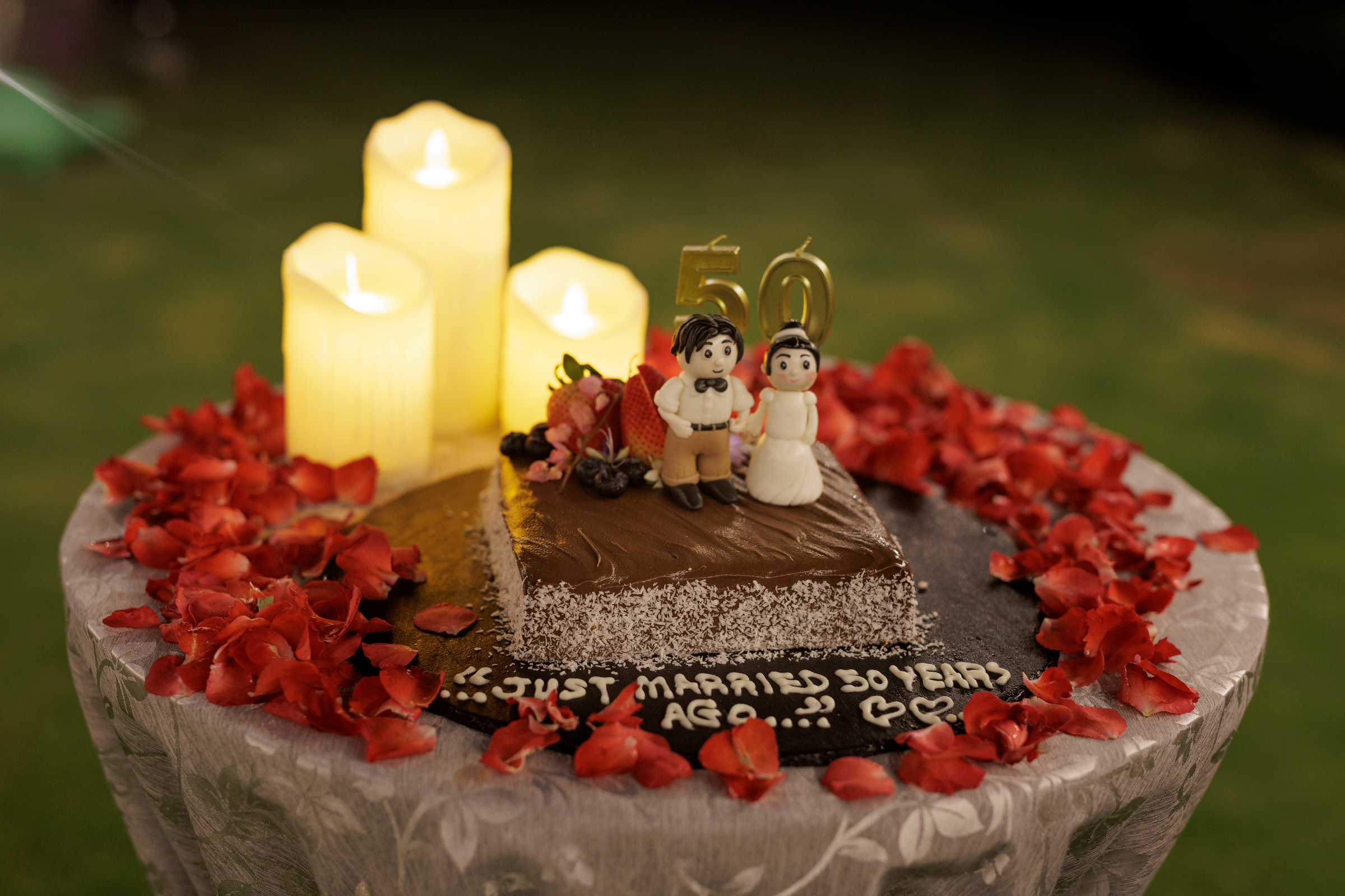 A decorated wedding anniversary cake with figurines of a bride and groom, surrounded by flower petals and candles, featuring the number 50.
