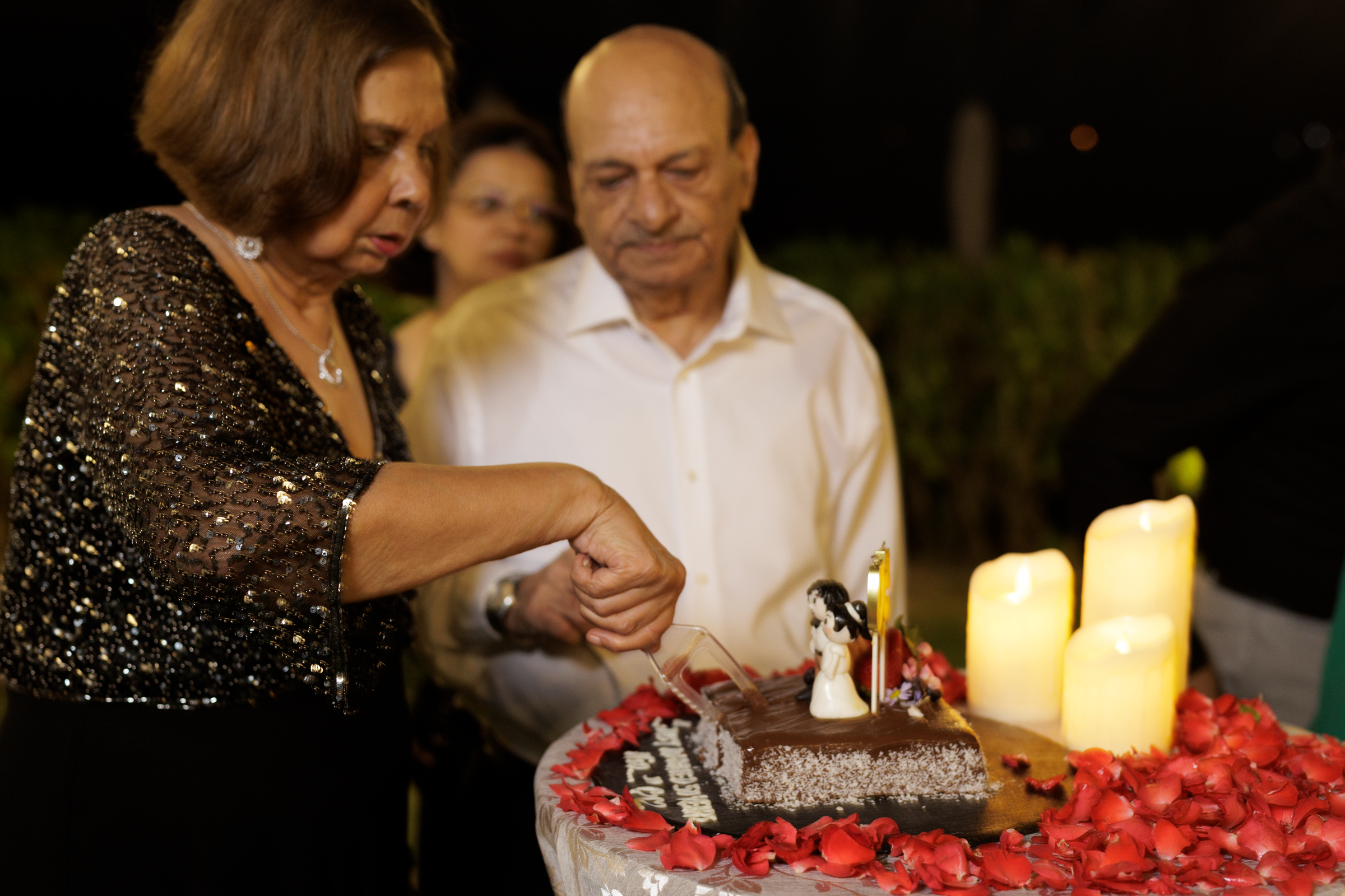 A woman in a sparkling black dress is cutting a birthday cake adorned with rose petals and candles, while a man observes attentively in a formal white shirt.