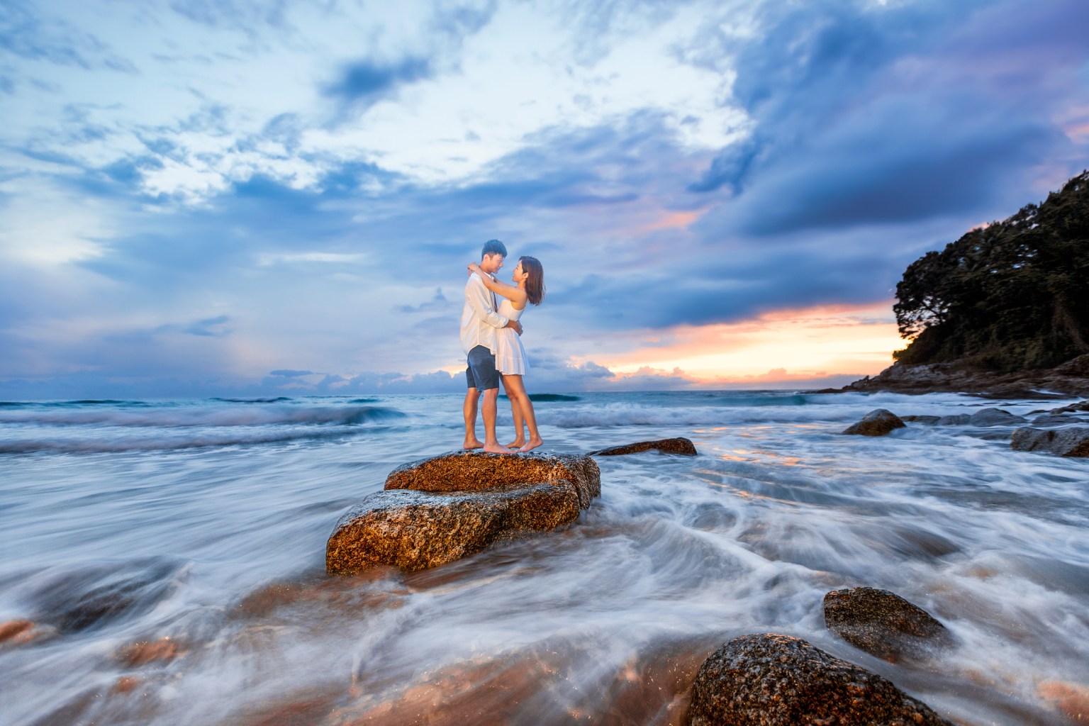 Couple embracing on a rock at Surin Beach during sunset, ocean waves gently splashing around them.
