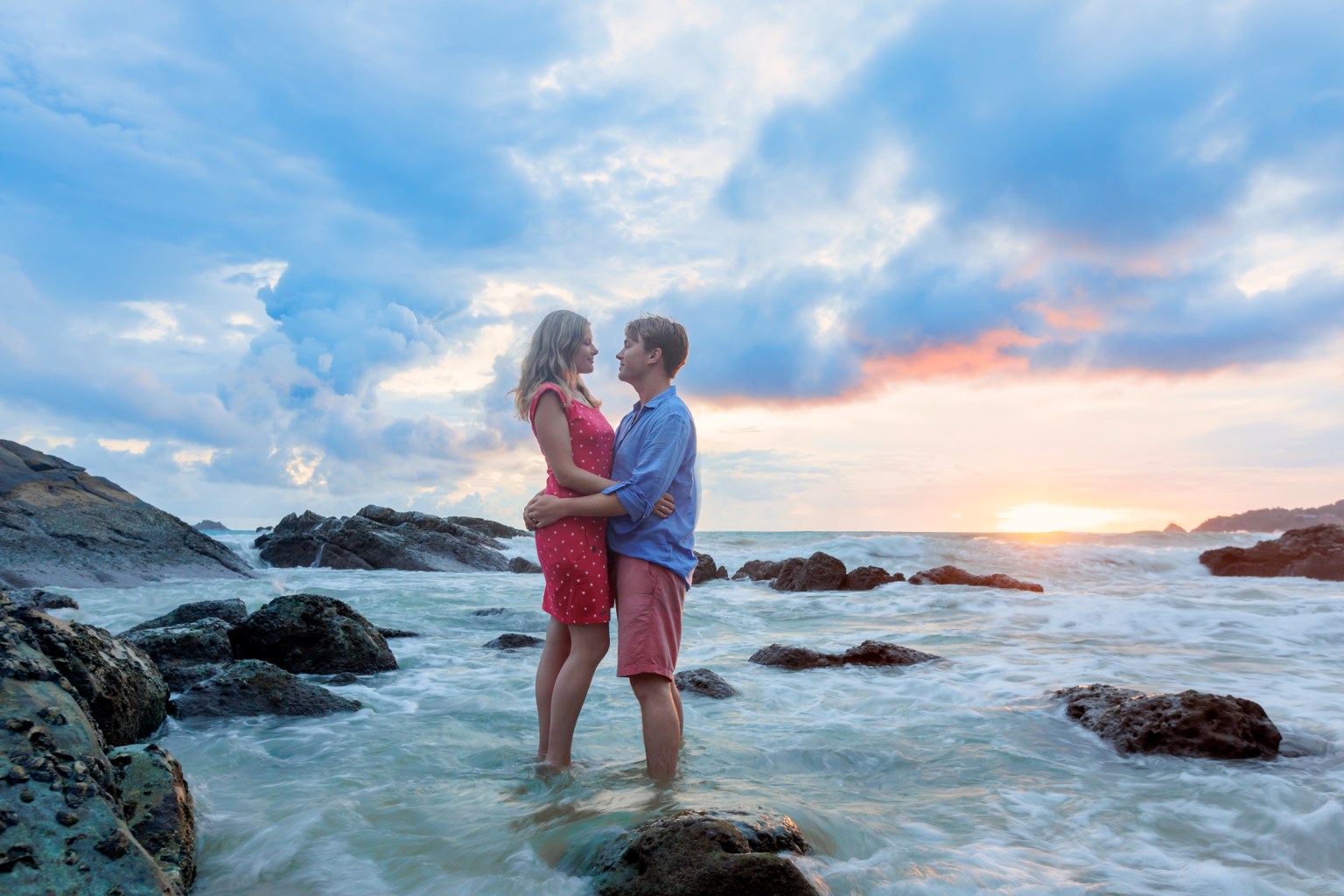 A couple standing on rocks in the ocean during sunset, with gentle waves around them and a colorful sky in the background at sea salt beach phuket