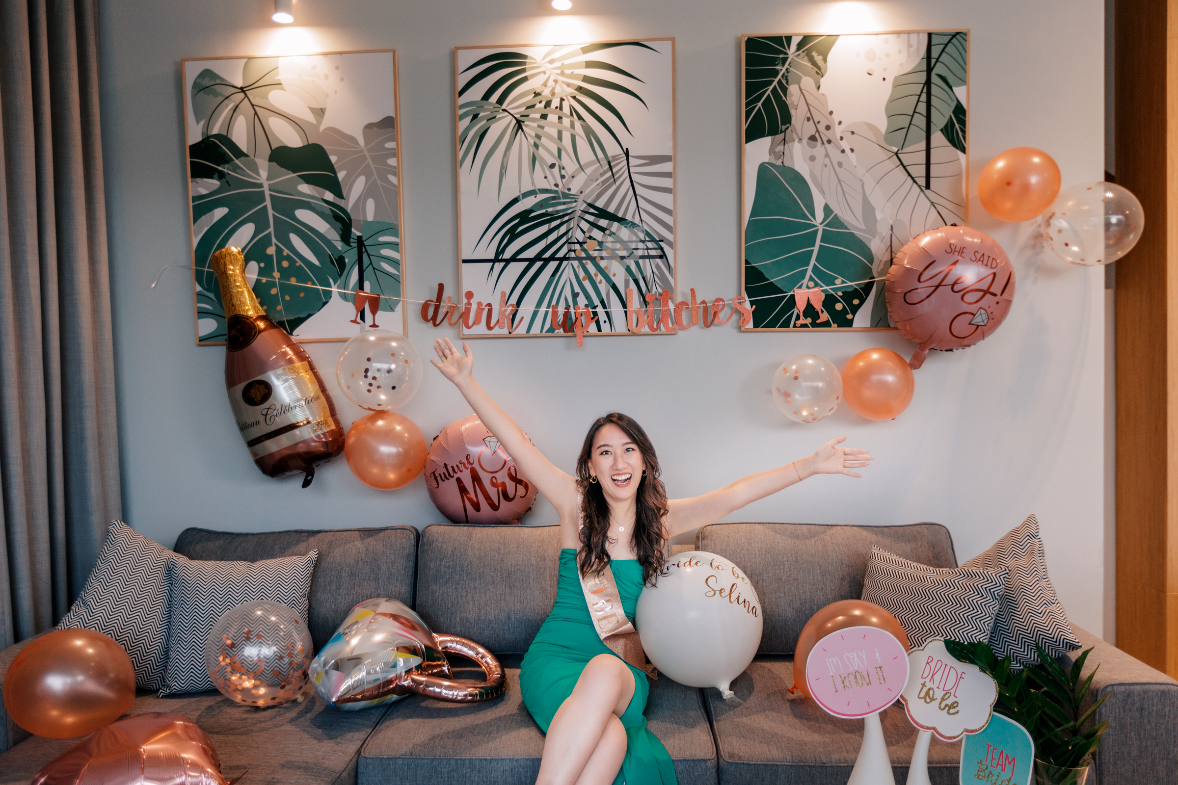 Bachelor party photography A woman wearing a green dress celebrating on a couch surrounded by balloons, decorations, and artwork featuring tropical plants.