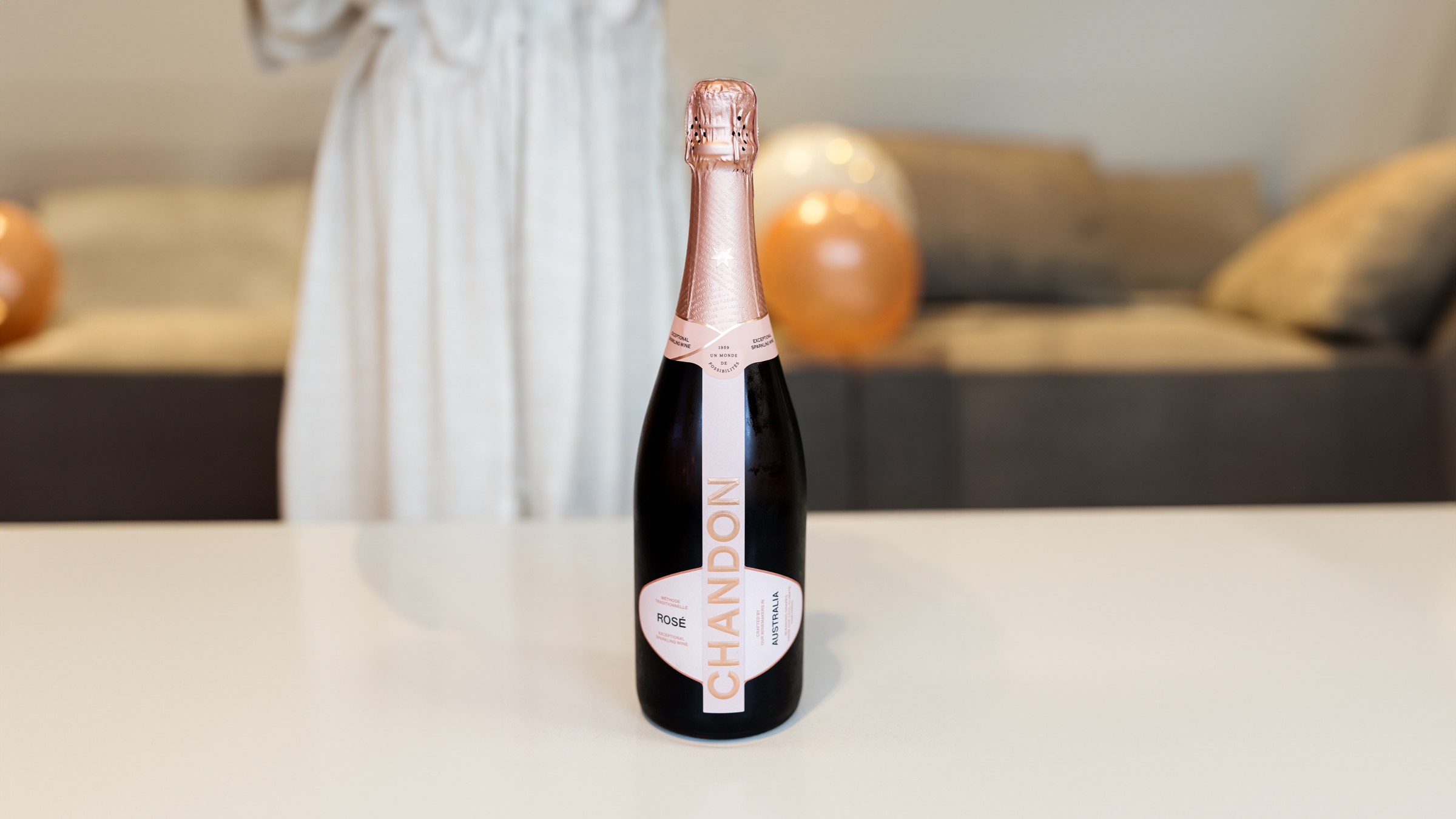 A bottle of sparkling wine stands on a table, with a person in a white dress blurred in the background and balloons nearby.