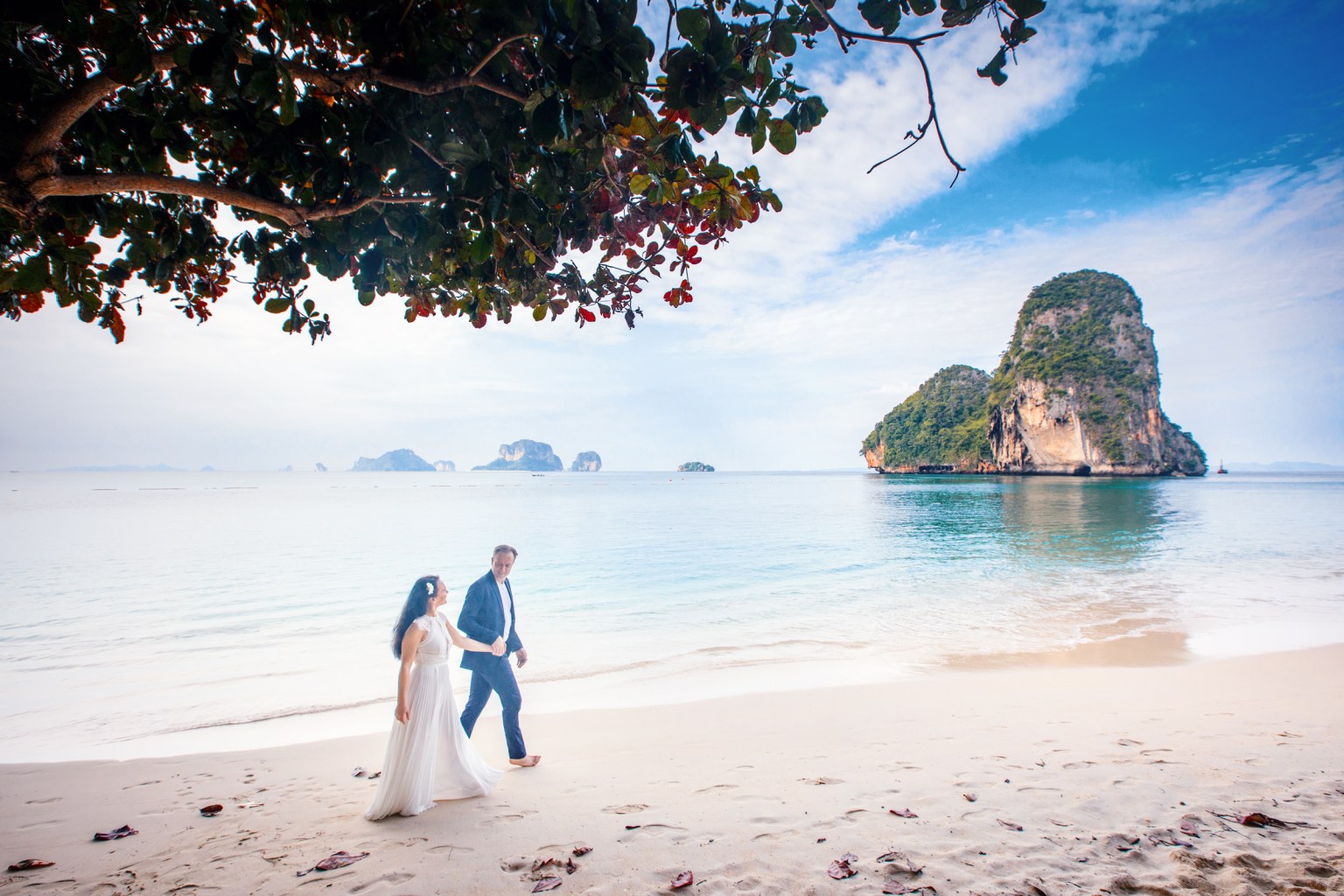 A couple walking hand in hand on a sandy beach in Krabi, Thailand, with limestone cliffs and islands in the background under a clear blue sky.